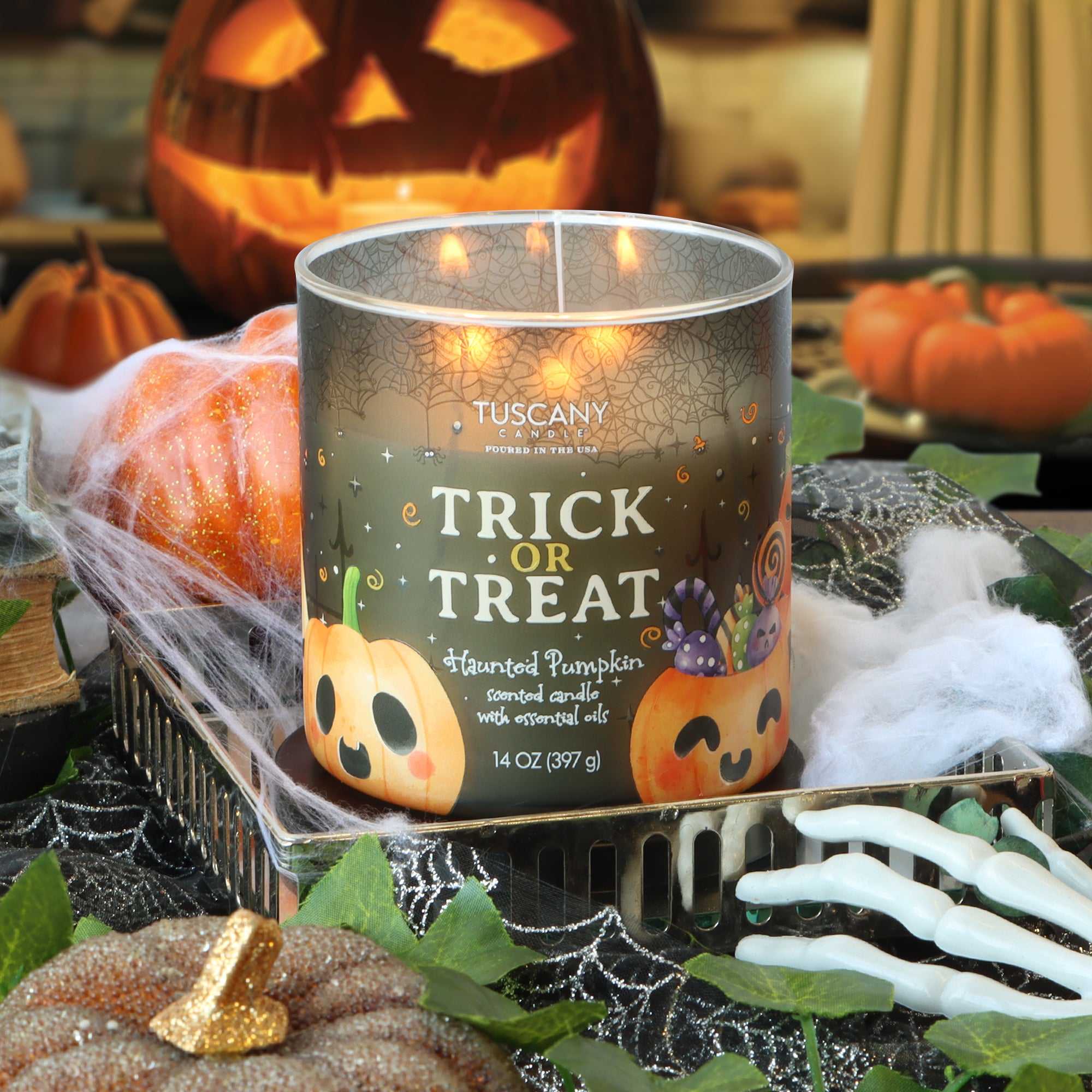 A Tuscany Candle® SEASONAL “Trick or Treat: Haunted Pumpkin Scented Halloween Candle” (14 oz) sits on a tray surrounded by Halloween decorations, including pumpkins, spider webs, and plastic bones.