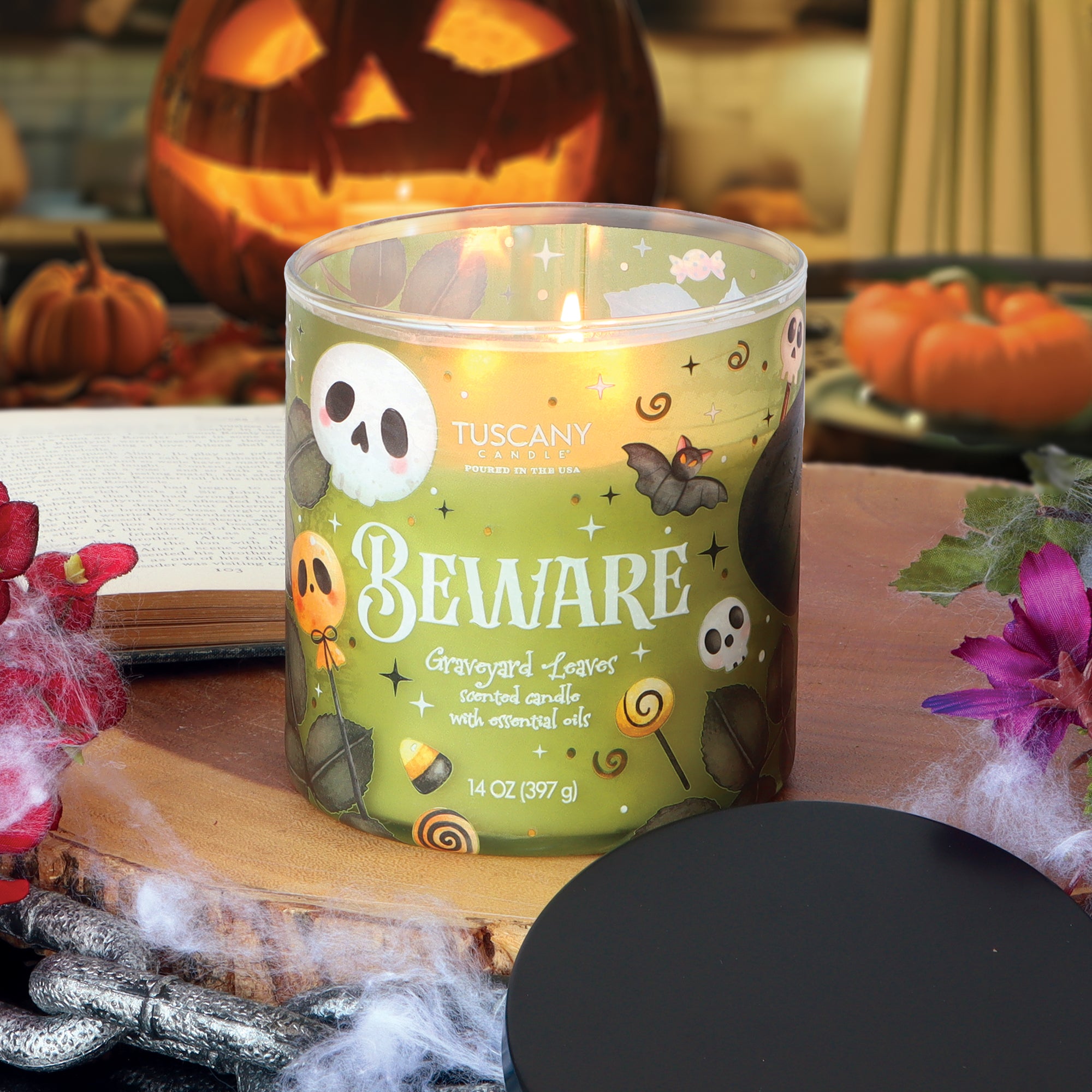 A 14 oz Tuscany Candle® SEASONAL “Beware: Graveyard Leaves Scented Halloween Candle,” featuring festive Halloween designs, sits on a table adorned with pumpkins, spider webs, and decorative flowers.