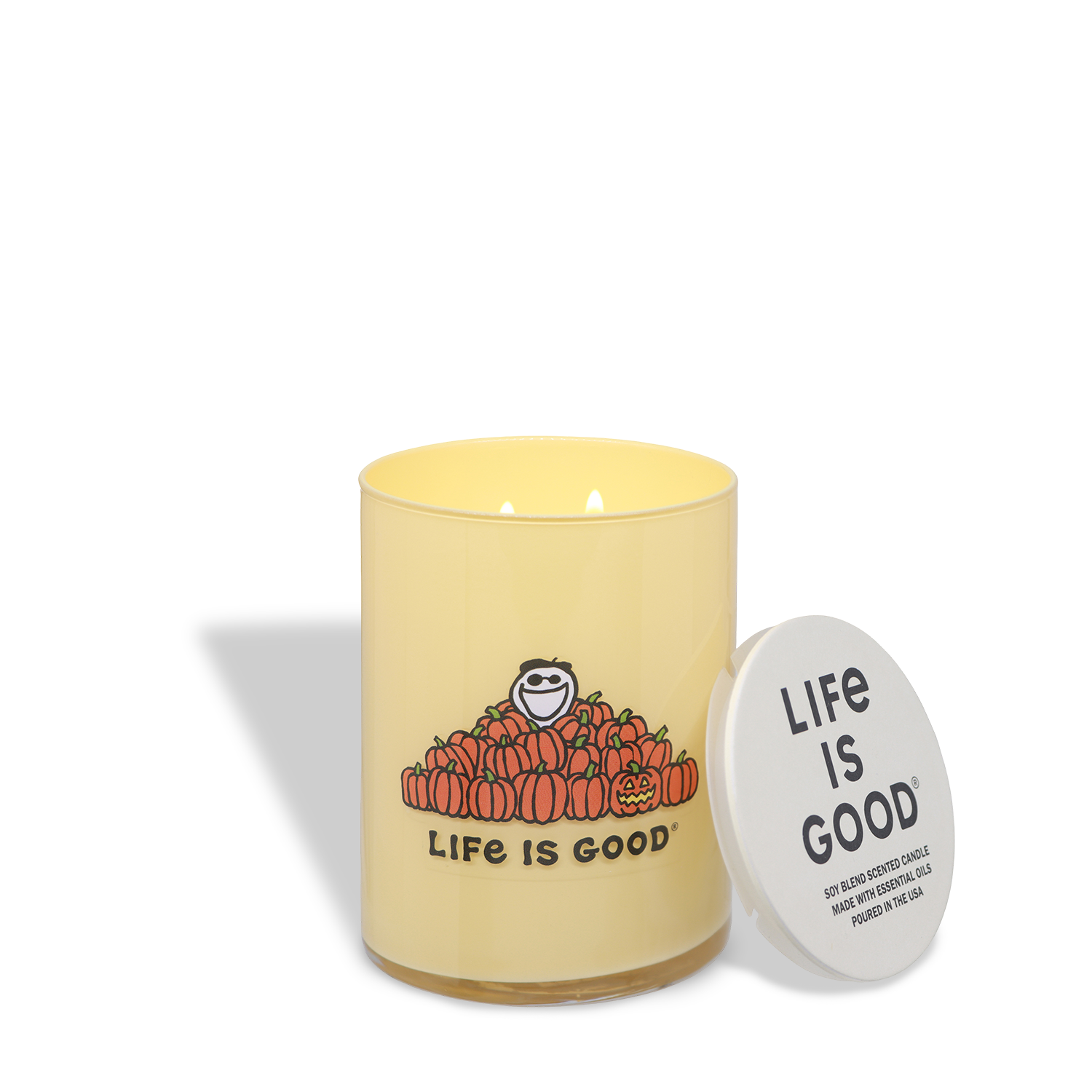 The Life Is Good: Vanilla Pumpkin Scented Halloween Candle (13 oz) by Tuscany Candle® SEASONAL features a yellow candle in a glass jar decorated with “Life Is Good” and a smiling figure surrounded by pumpkins; the white lid, printed with “Life Is Good,” is placed next to the candle.