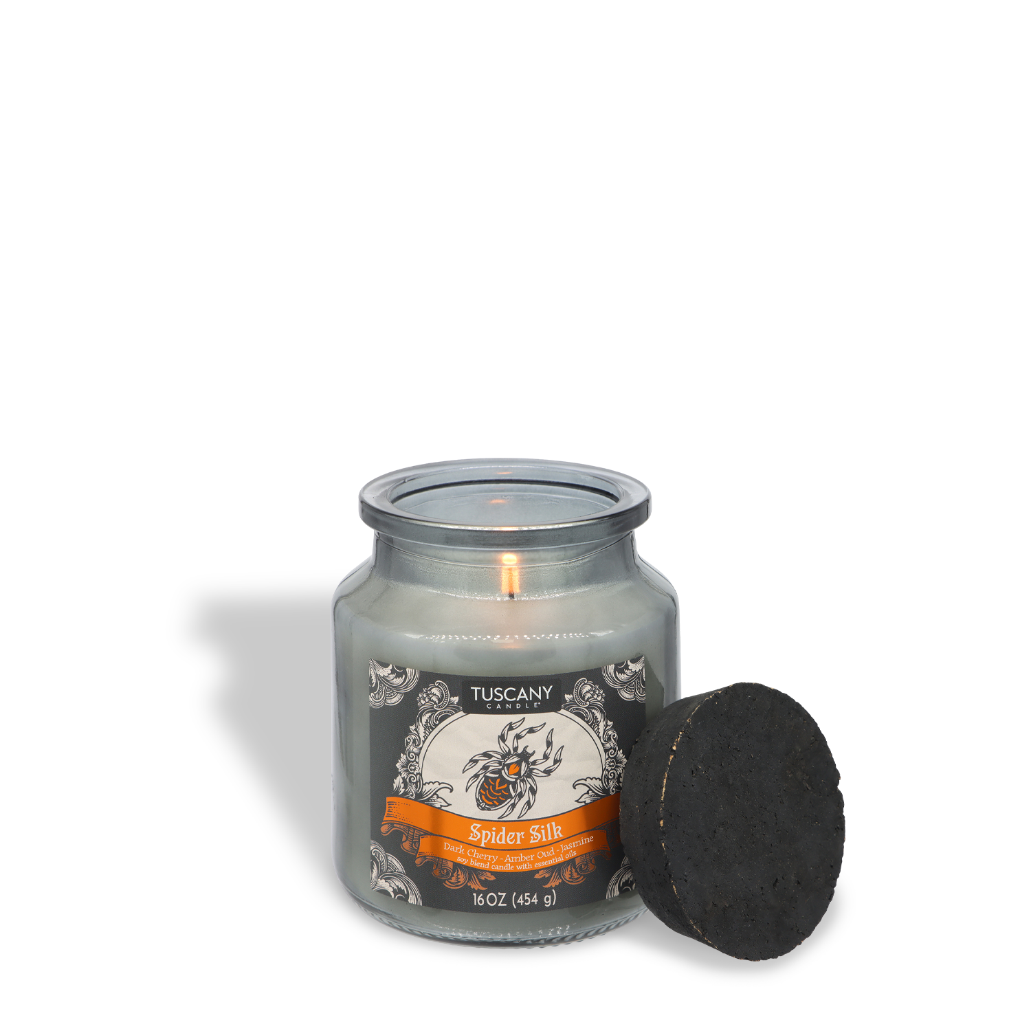 A lit white candle in a clear glass jar labeled “Spider Silk: Dark Cherry & Amber Oud” by Tuscany Candle® SEASONAL, with a black round lid resting next to it on a white background.