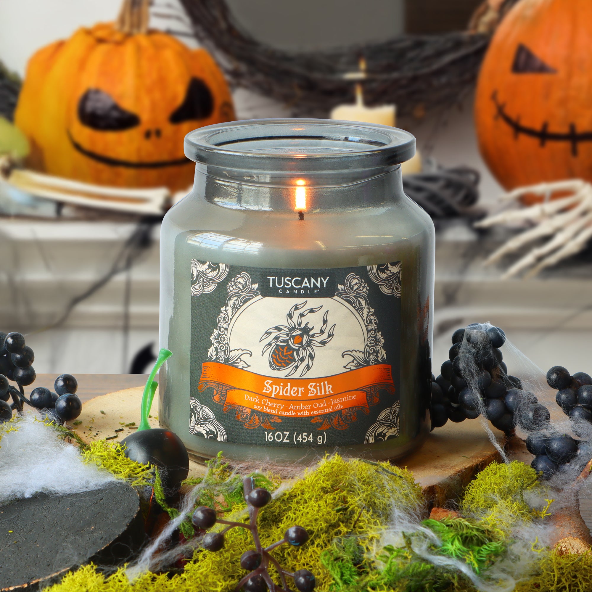 A Tuscany Candle® SEASONAL “Spider Silk: Dark Cherry & Amber Oud Scented Halloween Candle” (16 oz) burns on a table adorned with moss, faux spider webs, and black berries, while two carved pumpkins can be seen in the background.