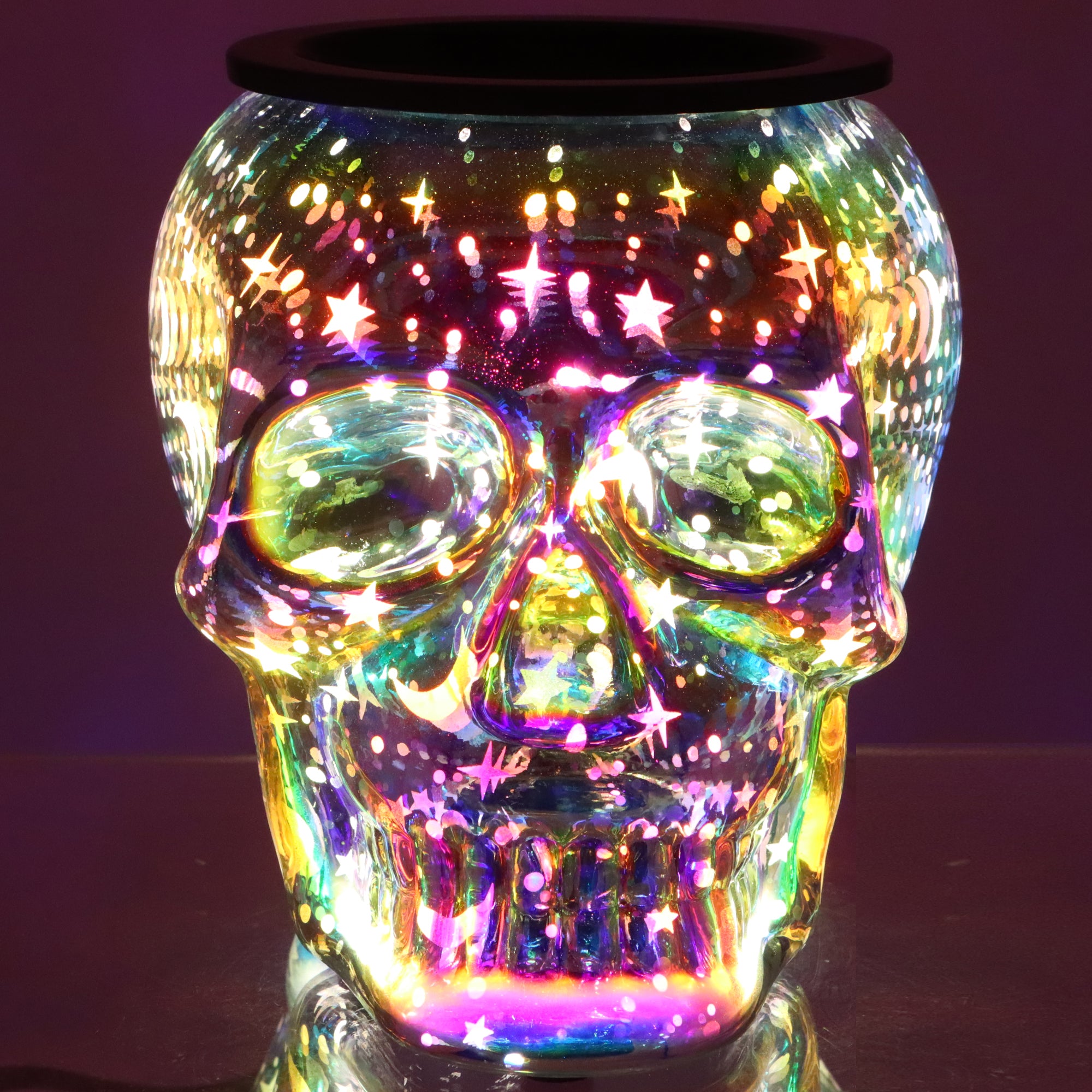 The Tuscany Candle® SEASONAL Holographic Skull Wax Melt Warmer features a glass skull design that emits multicolored light patterns with star and dot motifs against a dark background.