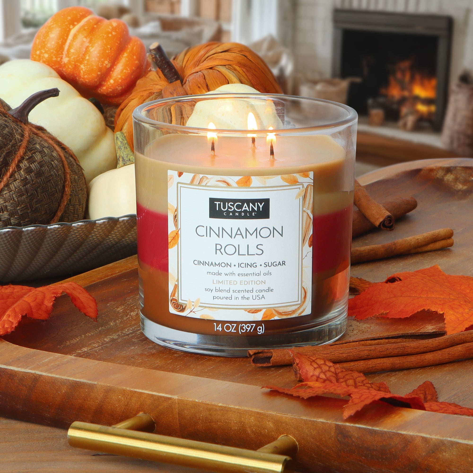A Tuscany Candle® SEASONAL Cinnamon Rolls: Icing & Sugar Scented Fall Candle (14 oz) glows on a wooden tray with autumn leaves, pumpkins, and cinnamon sticks, as a fireplace and decorative gourds set a cozy backdrop.