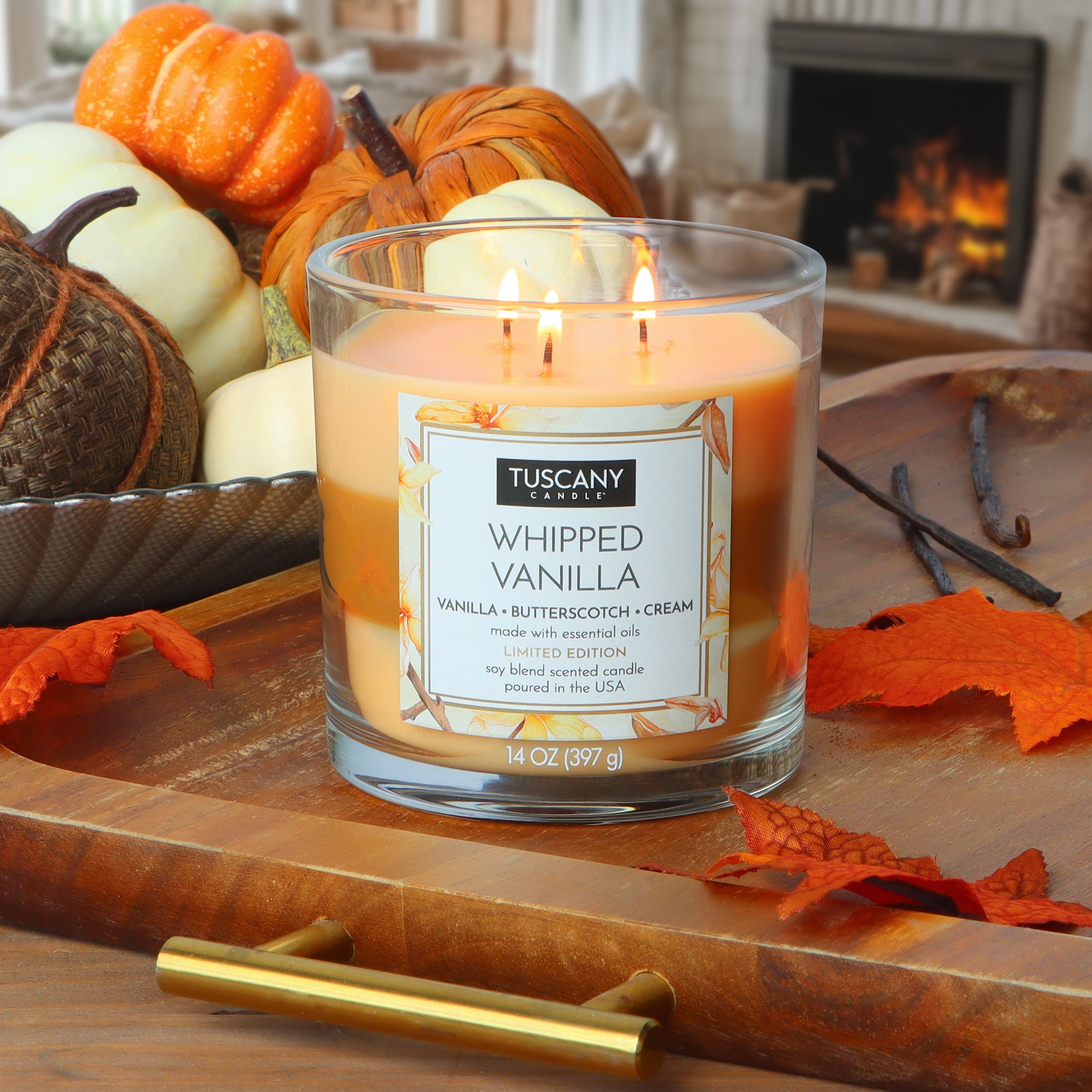 A Tuscany Candle® SEASONAL Whipped Vanilla: Butterscotch & Cream Scented Fall Candle (14 oz) burns on a wooden tray with orange leaves, surrounded by fall décor and a lit fireplace in the background.