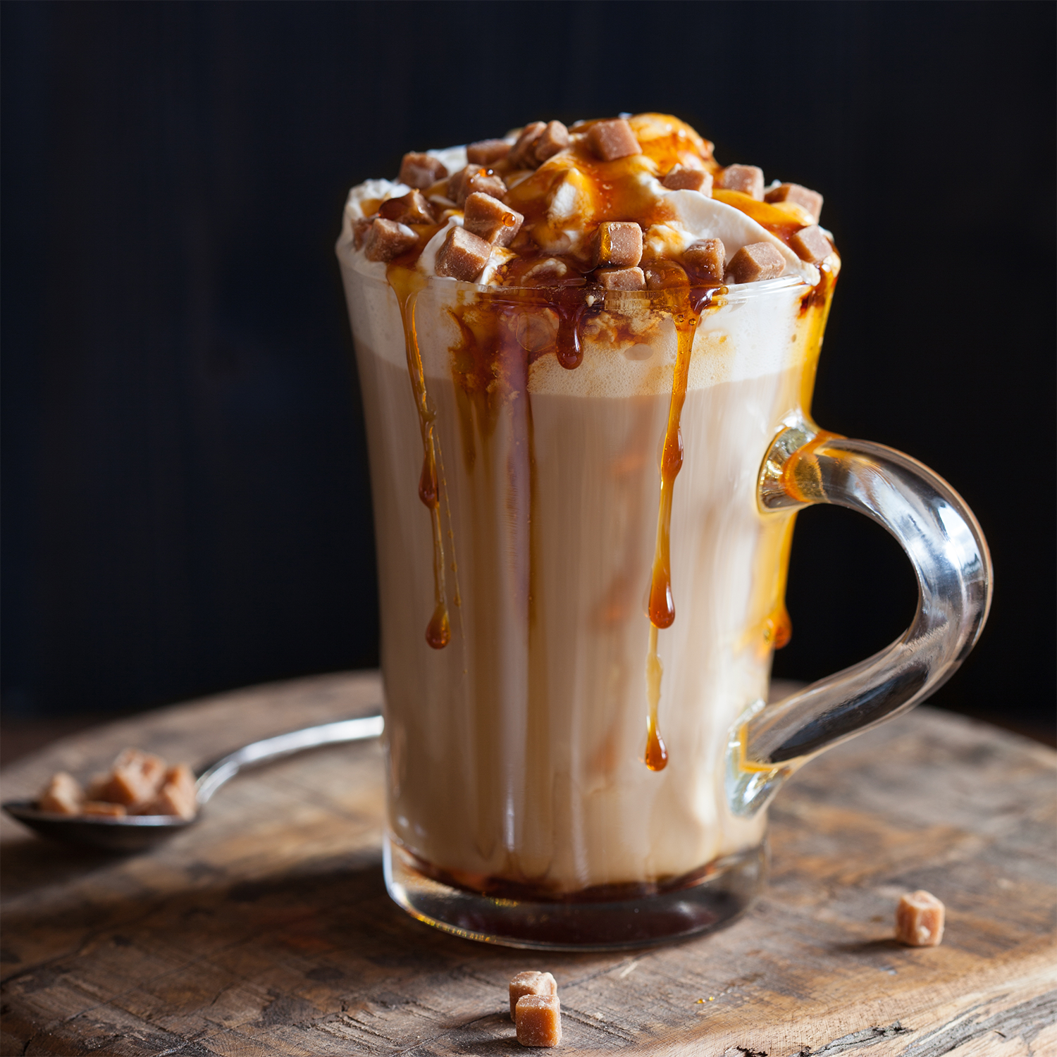Nestled on a wooden surface sits a glass mug of caramel latte, crowned with whipped cream, luscious caramel drizzle, and chunks of caramel—with extra pieces resting on a nearby spoon—creating an inviting scene as warm and cozy as the comforting glow and rich aroma of Tuscany Candle® SEASONAL's Whipped Vanilla: Butterscotch & Cream Scented Fall Candle (14 oz) filling your home.