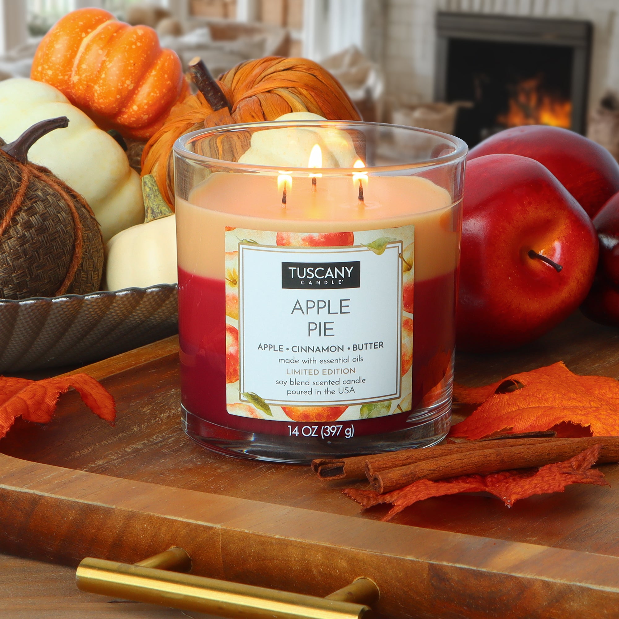 A lit Tuscany Candle® SEASONAL "Apple Pie: Cinnamon & Butter Scented Fall Candle" (14 oz) sits on a wooden tray with autumn leaves, apples, and pumpkins, backed by a fireplace and armchair.