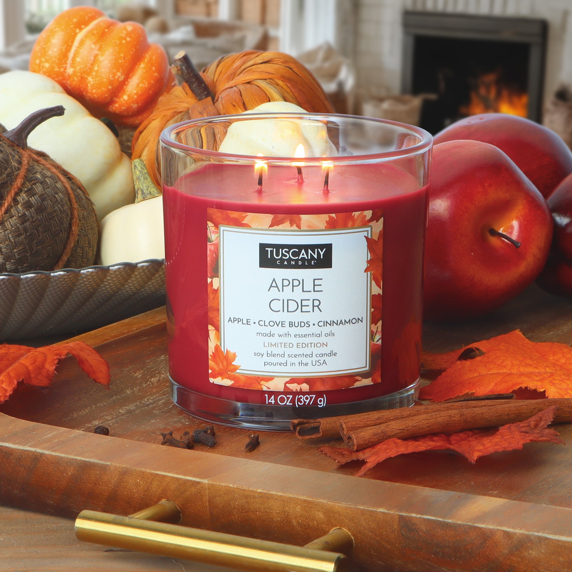 A Tuscany Candle® SEASONAL Apple Cider: Clove Buds & Cinnamon Scented Fall Candle (14 oz) glows on a wooden table with pumpkins, apples, and autumn leaves nearby, while a fireplace flickers in the background.