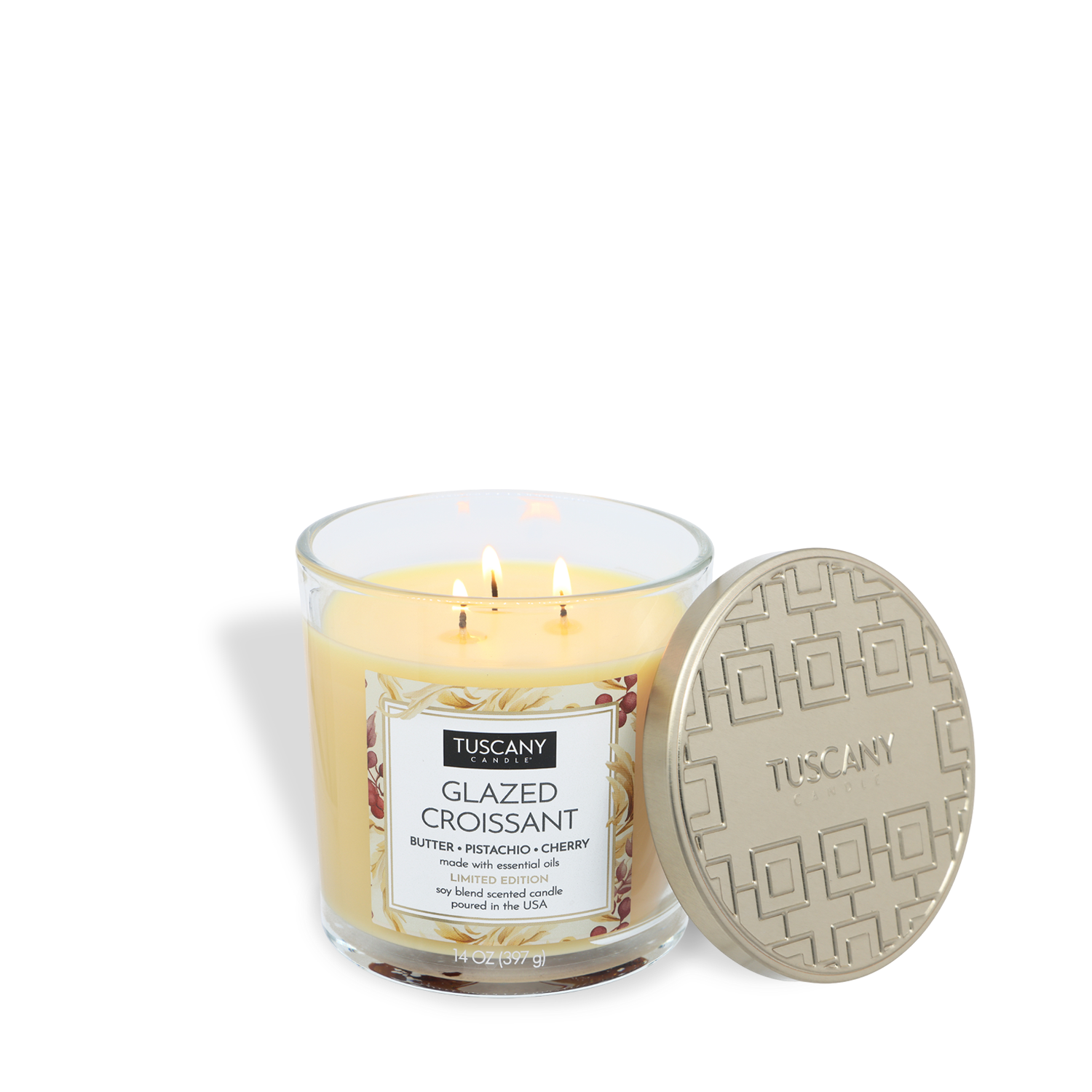A Tuscany Candle® SEASONAL "Glazed Croissant: Butter & Pistachio Scented Fall Candle" (14 oz) with three lit wicks sits beside a beige patterned lid embossed with "Tuscany.