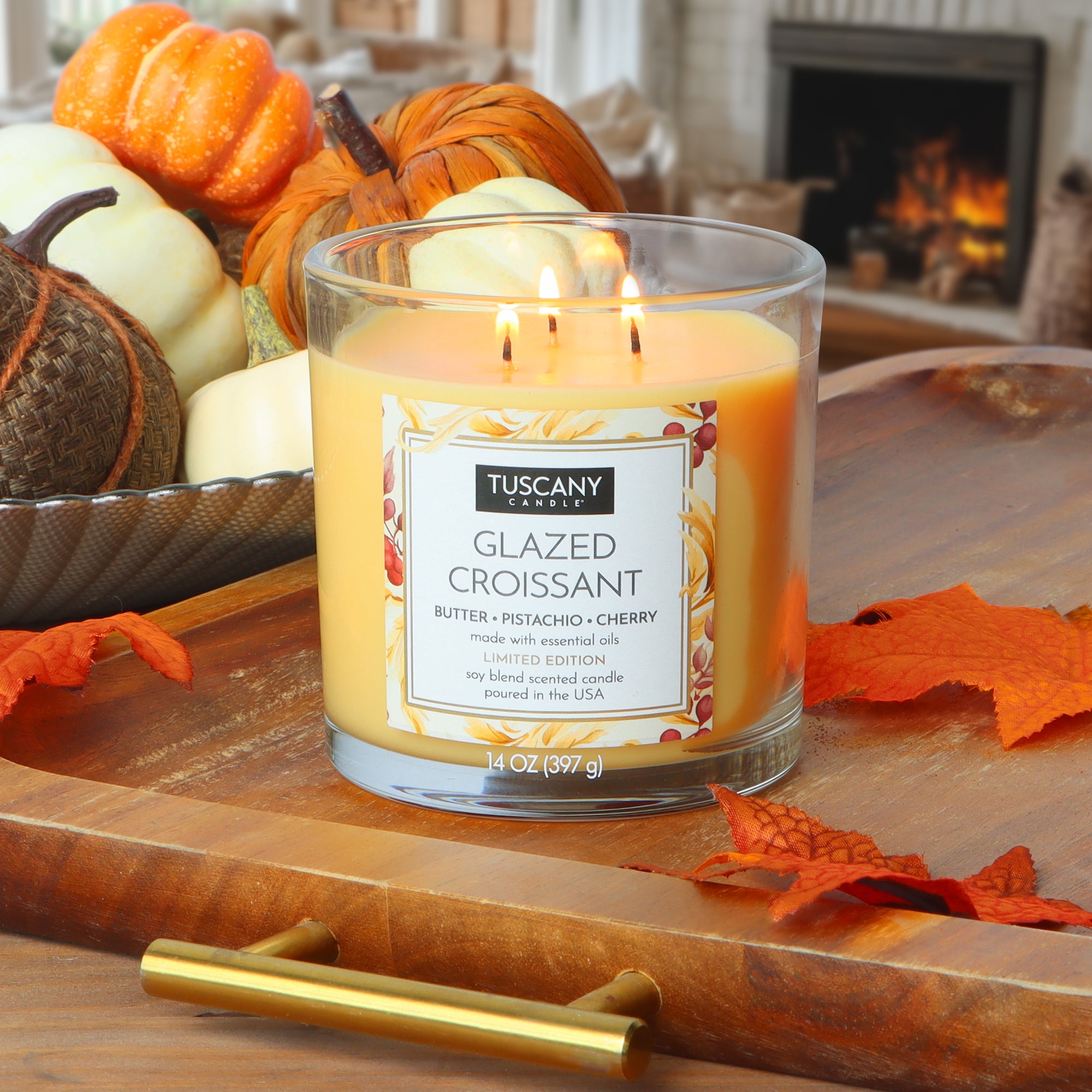 A Tuscany Candle® SEASONAL "Glazed Croissant: Butter & Pistachio Scented Fall Candle" (14 oz, two wicks) glows on a wooden tray amid pumpkins and autumn leaves, creating a cozy indoor vibe.