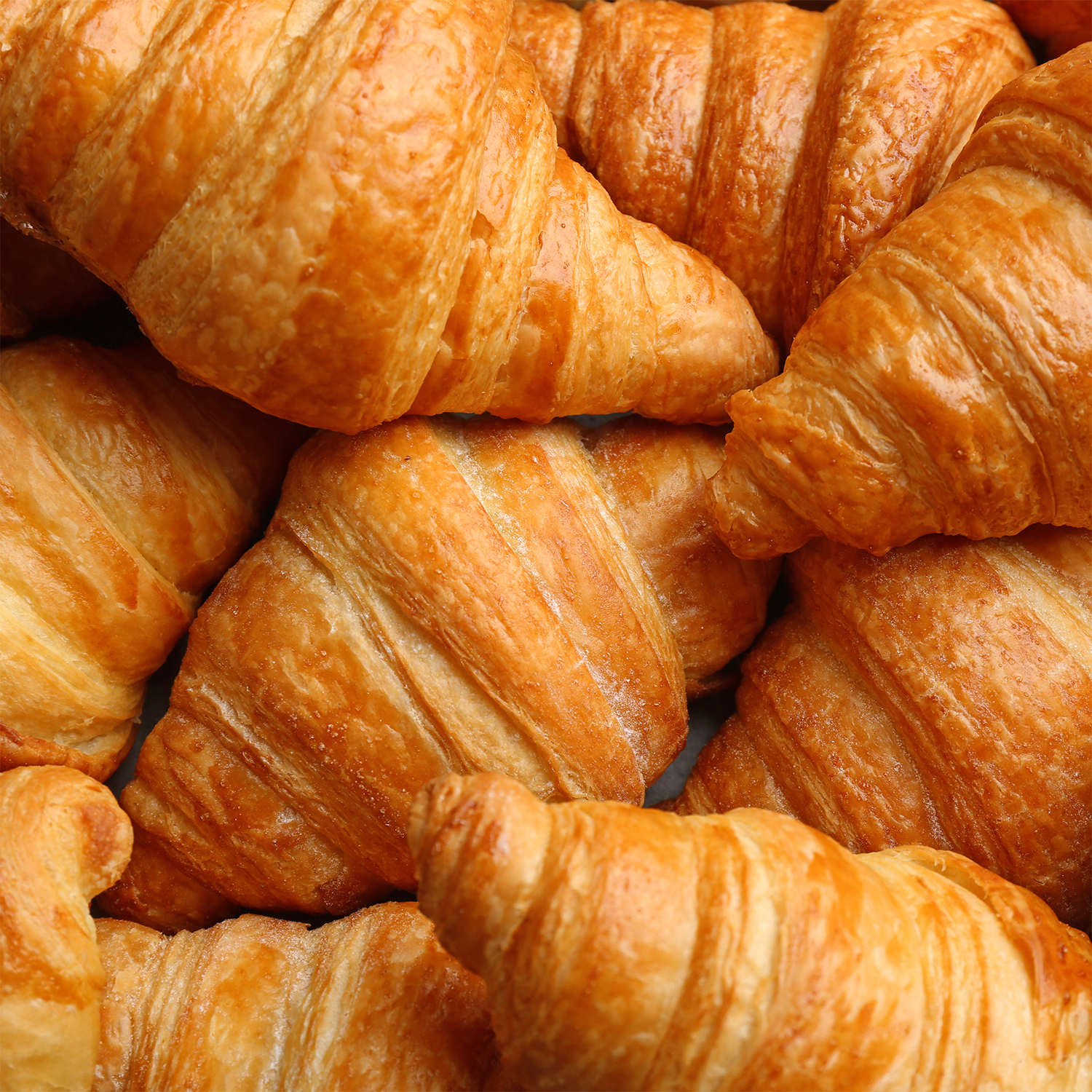 A close-up view of several golden-brown croissants stacked together, highlighting their flaky, layered texture—evoking the comforting bakery-inspired aroma of Tuscany Candle® SEASONAL's Glazed Croissant: Butter & Pistachio Scented Fall Candle (14 oz), which fills the air with a warm, autumnal fragrance.
