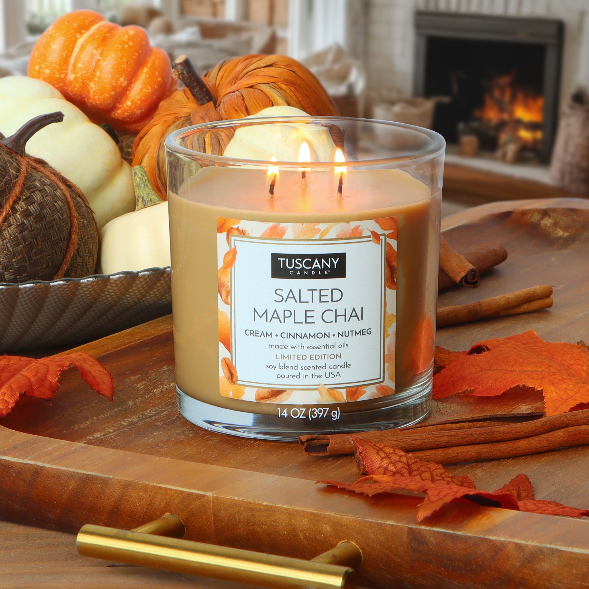 A Tuscany Candle® SEASONAL Salted Maple Chai: Cream & Cinnamon Scented Fall Candle (14 oz) glows on a wooden tray surrounded by cinnamon sticks, small pumpkins, and autumn leaves, with a fireplace in the background.