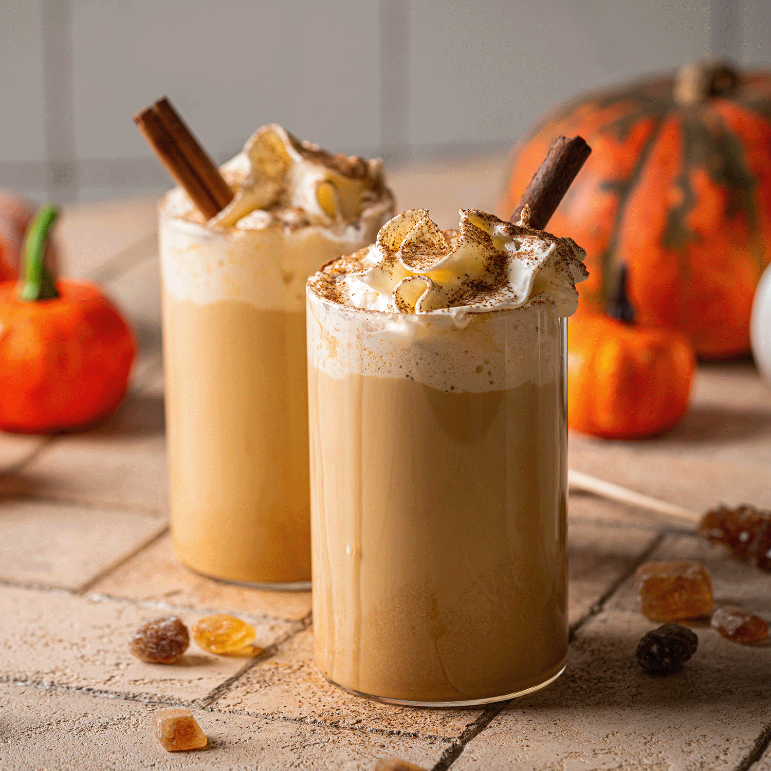 Two jars of Tuscany Candle® SEASONAL's Salted Maple Chai: Cream & Cinnamon Scented Fall Candle (14 oz) sit on a tiled surface, their comforting fragrance evoking the essence of autumn with creamy chai and warm cinnamon notes, while small pumpkins in the background add a cozy, seasonal touch.