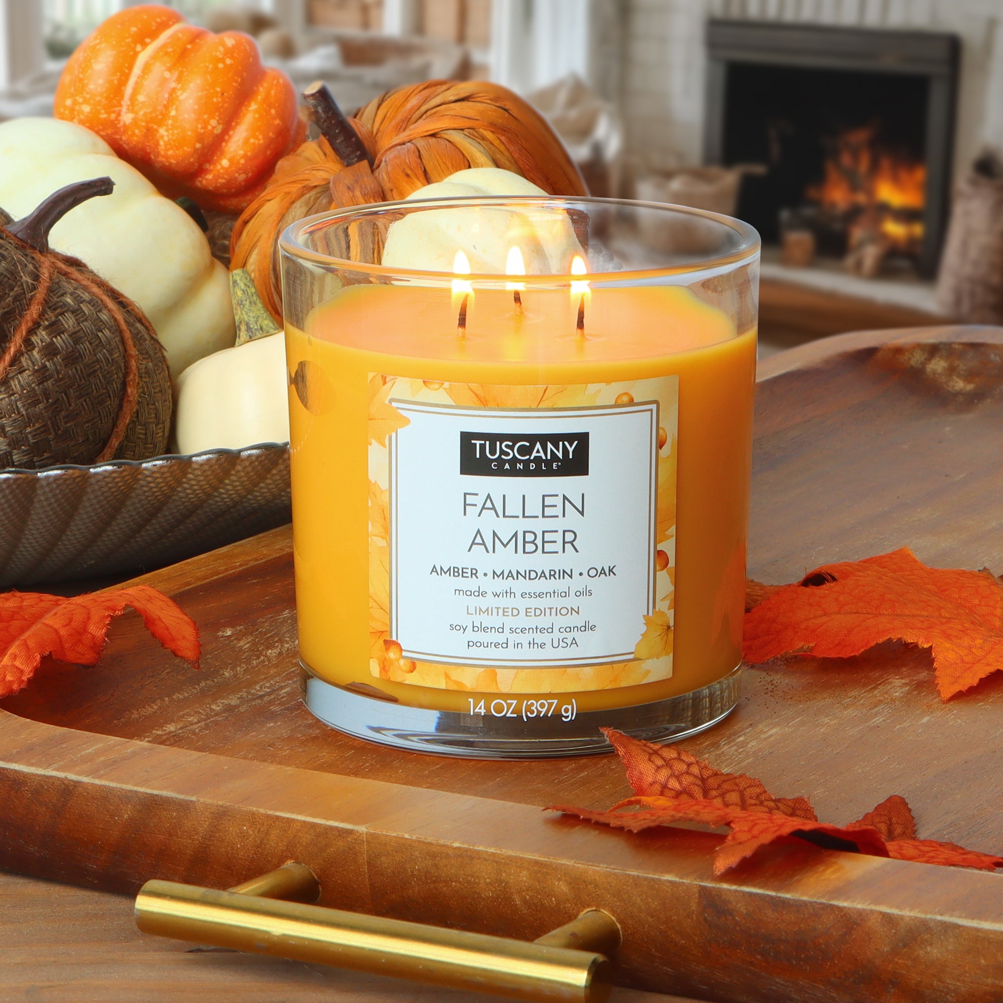 A lit Tuscany Candle® SEASONAL Fallen Amber: Mandarin & Oak Scented Fall Candle (14 oz) sits on a wooden tray with autumn leaves, small pumpkins, and a fireplace in the background.