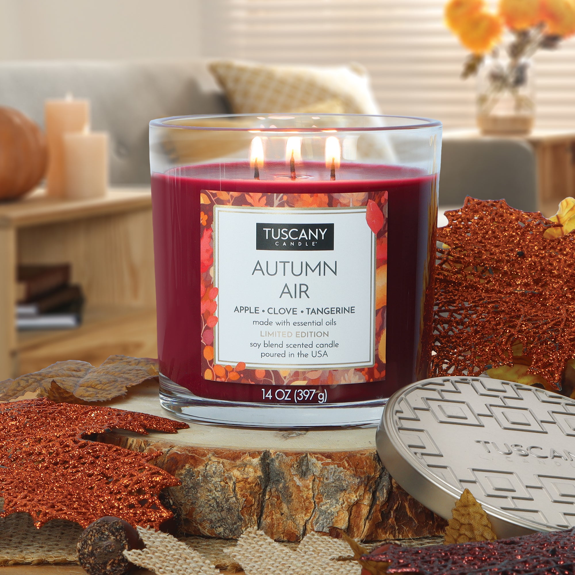 A red, three-wick Tuscany Candle® SEASONAL "Autumn Air: Apple & Clove Scented Fall Candle" (14 oz) sits on a wooden surface with autumn leaves and seasonal decor in the background.