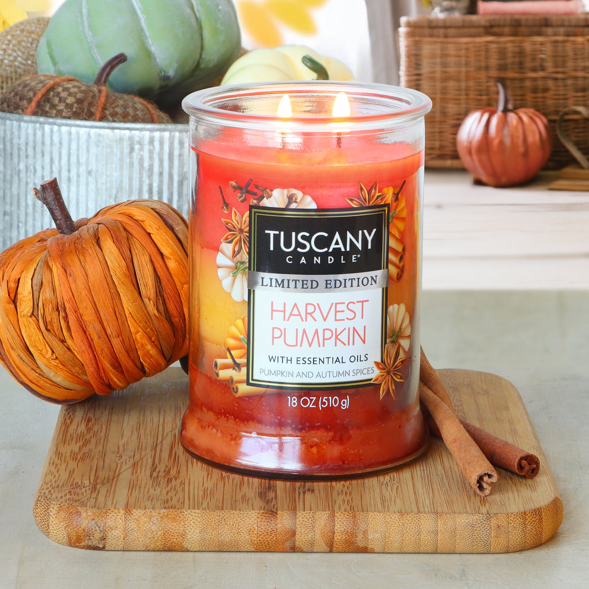 A lit Tuscany Candle® SEASONAL "Harvest Pumpkin: Pumpkin & Autumn Spices Scented Fall Candle (18 oz)" sits on a wooden board beside cinnamon sticks, with autumn decor and pumpkins in the background.