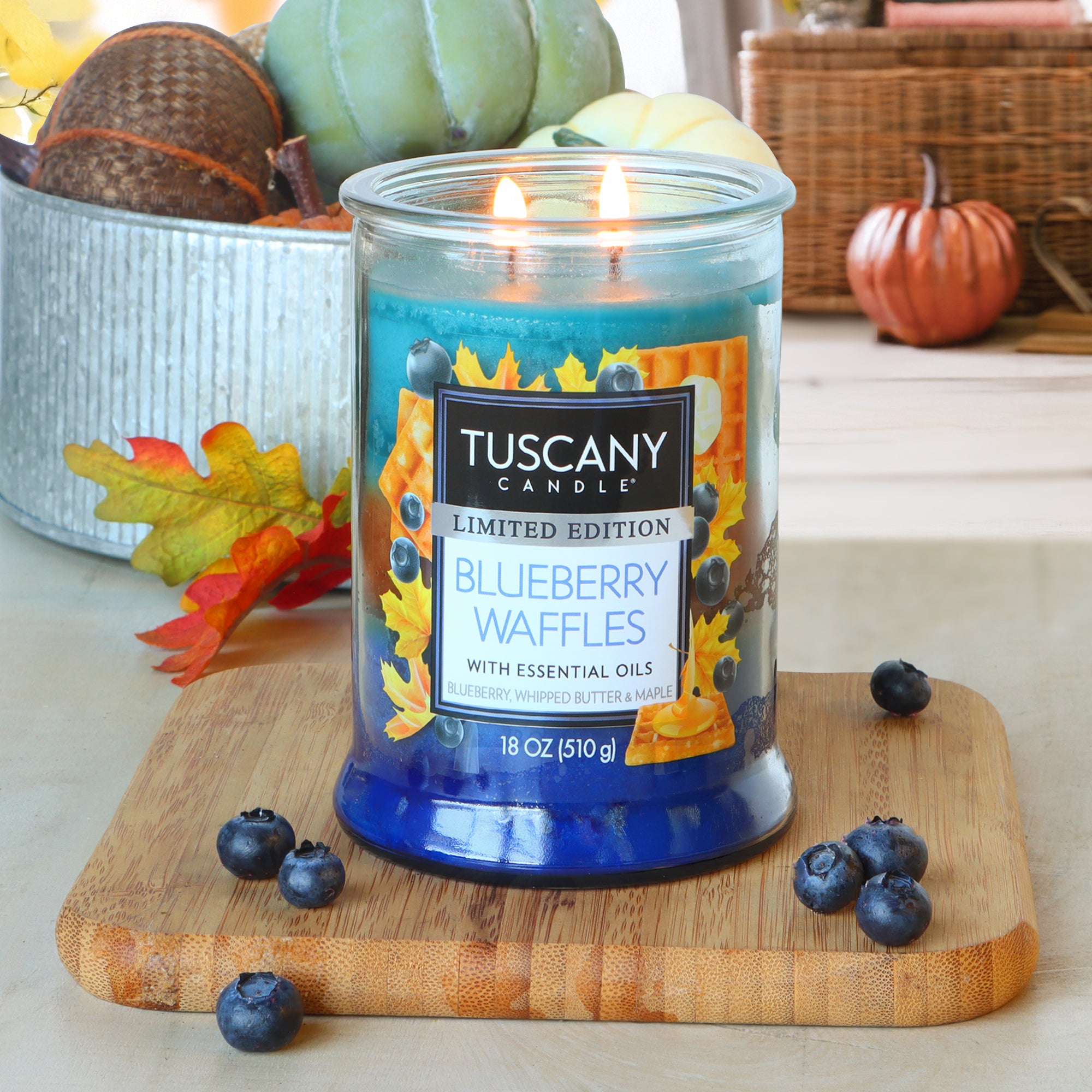 An 18 oz Tuscany Candle® SEASONAL "Blueberry Waffles: Whipped Butter & Maple Scented Fall Candle" sits on a wooden board, lit with both wicks and surrounded by blueberries and autumn decor.
