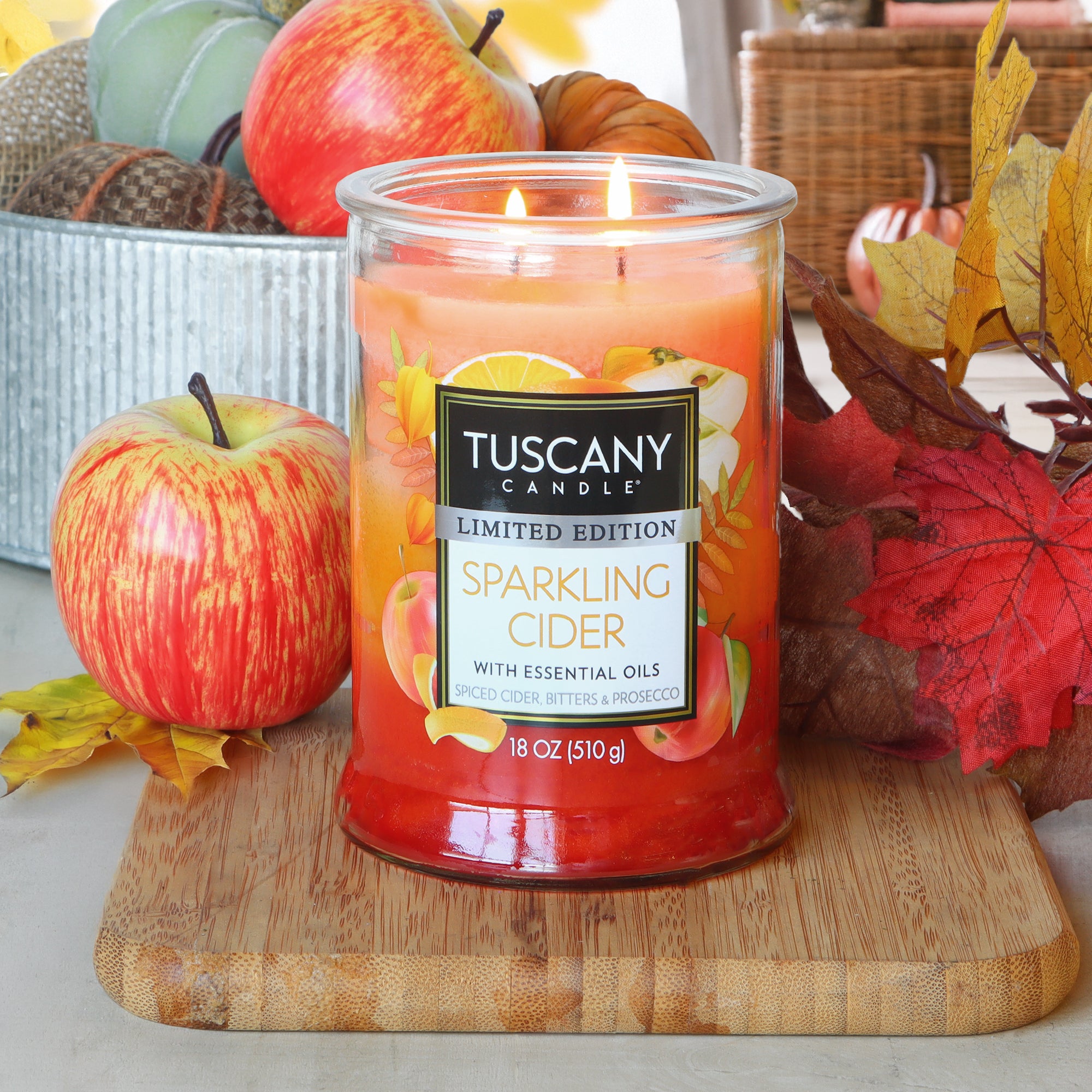 A lit Tuscany Candle® SEASONAL "Sparkling Cider: Bitters & Prosecco Scented Fall Candle" (18 oz) rests on a wooden board, surrounded by artificial apples, autumn leaves, and festive fall decor.
