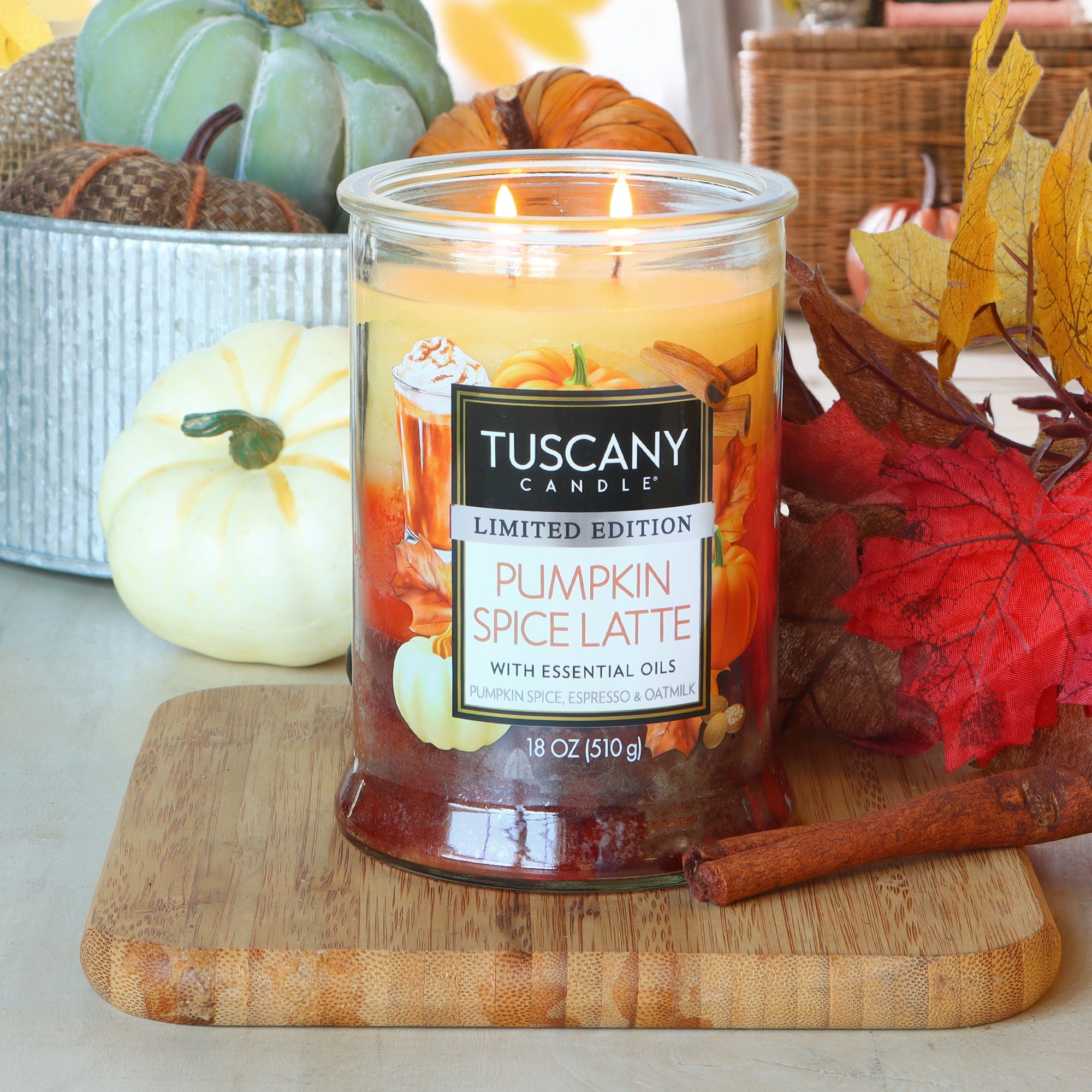 A Tuscany Candle® SEASONAL Pumpkin Spice Latte: Espresso & Oat Milk Scented Fall Candle (18 oz) glows on a wooden board surrounded by small pumpkins, autumn leaves, and cinnamon sticks.