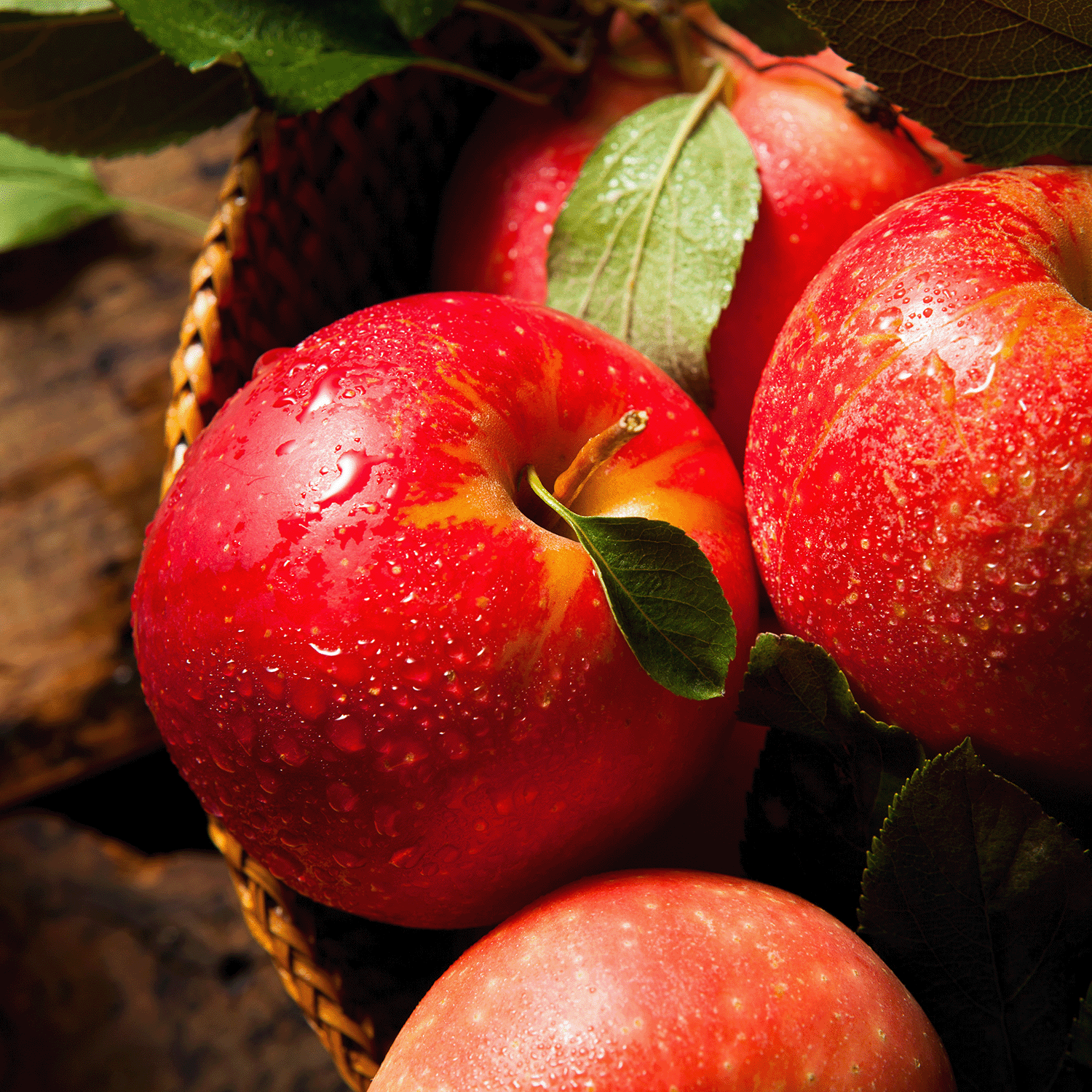 Close-up of fresh red apples with water droplets in a woven basket, surrounded by green leaves on a wooden surface—an inviting scene that captures the comforting aroma of Hickory Apple: Spiced Apple & Golden Hay, an 18 oz scented fall candle from Tuscany Candle® SEASONAL, evoking the cozy warmth of an autumn home fragrance.