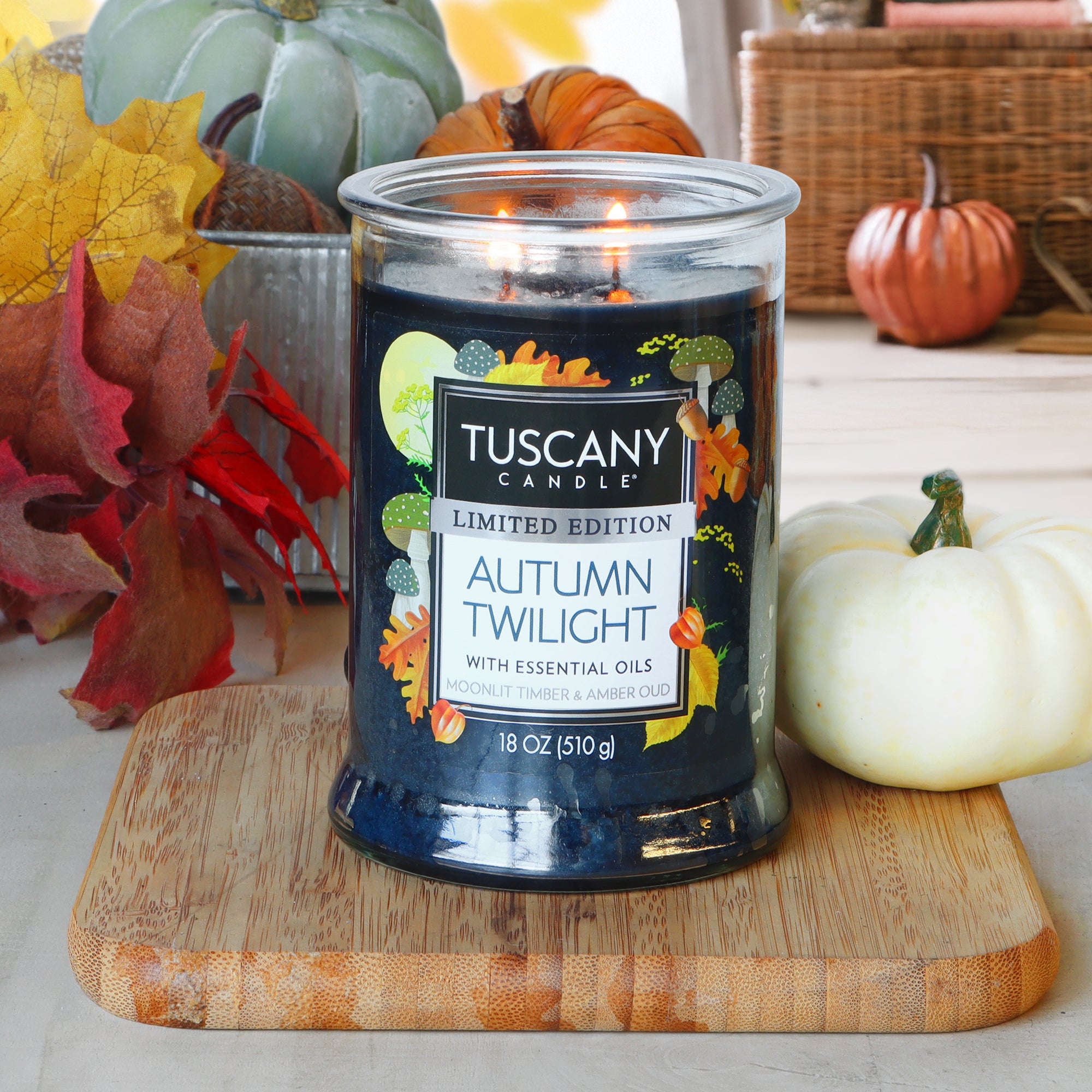 An 18 oz Tuscany Candle® SEASONAL, "Autumn Twilight: Moonlit Timber & Amber Oud," sits lit on a wooden board, surrounded by artificial pumpkins and autumn leaves.