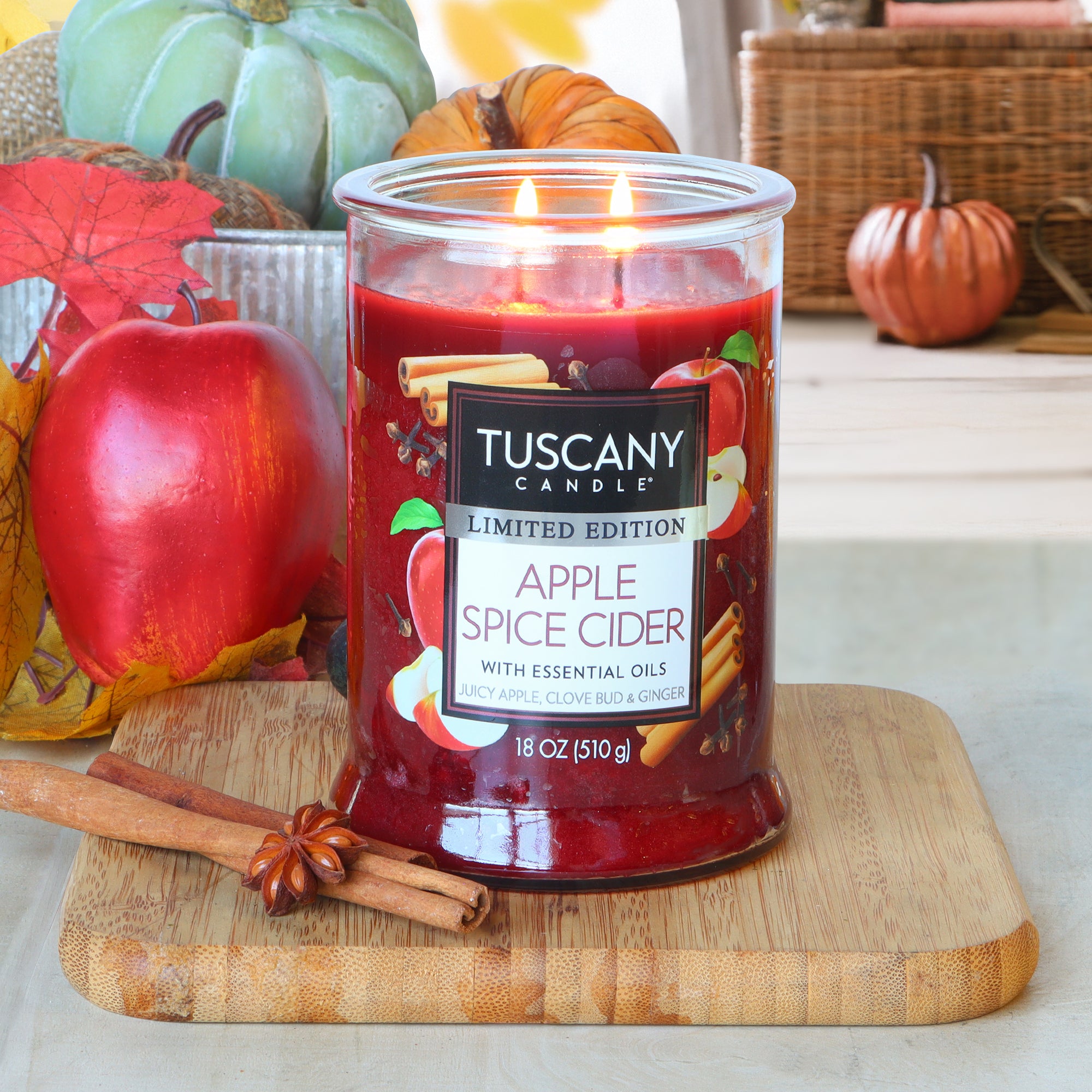 A lit Tuscany Candle® SEASONAL Apple Spice Cider: Clove Bud & Ginger scented fall candle (18 oz) sits on a wooden board with an apple, cinnamon sticks, and autumn decor in the background.