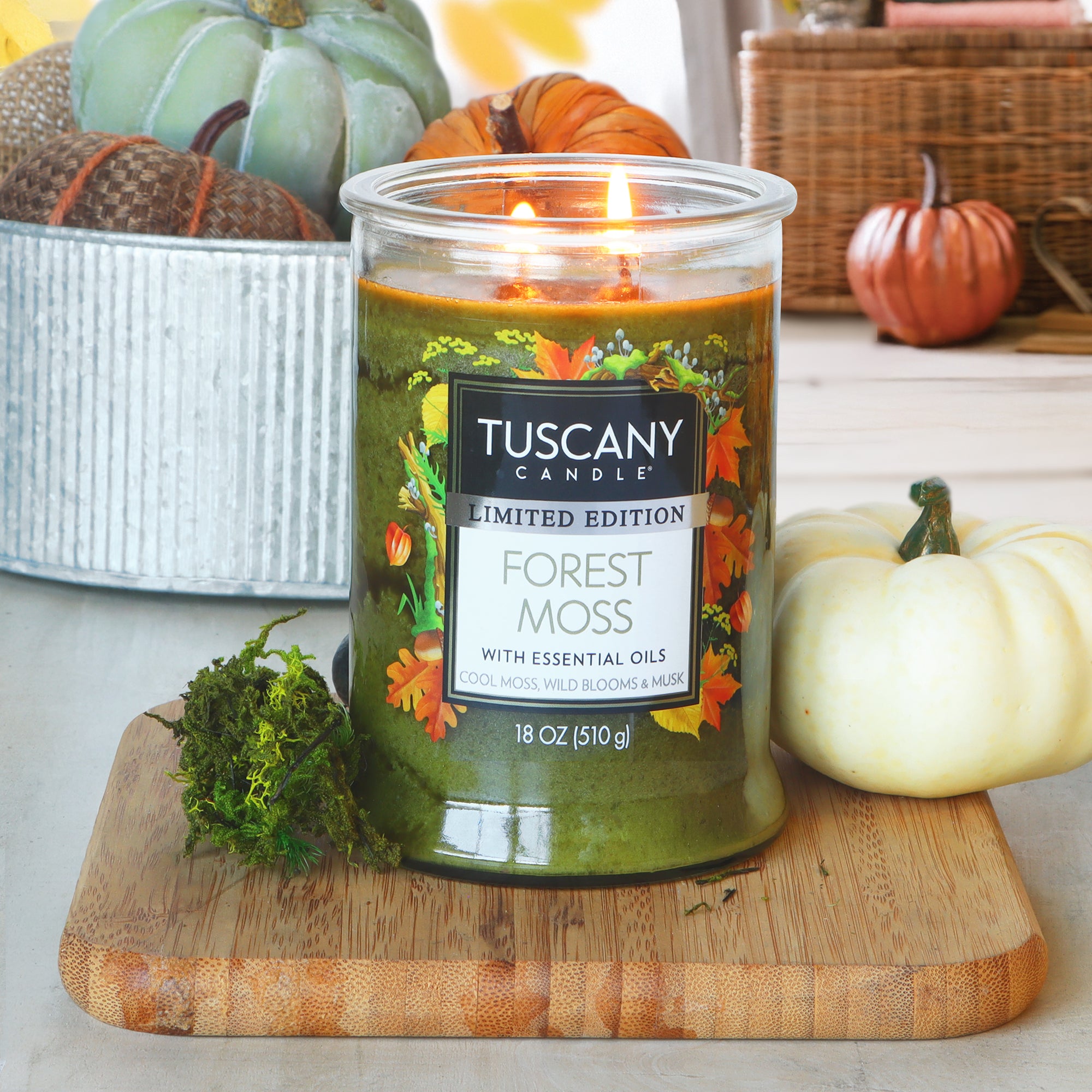A lit Tuscany Candle® SEASONAL "Forest Moss: Wild Blooms & Musk" 18 oz fall candle is on a wooden board with moss and a small white pumpkin, accented by autumn décor in the background.