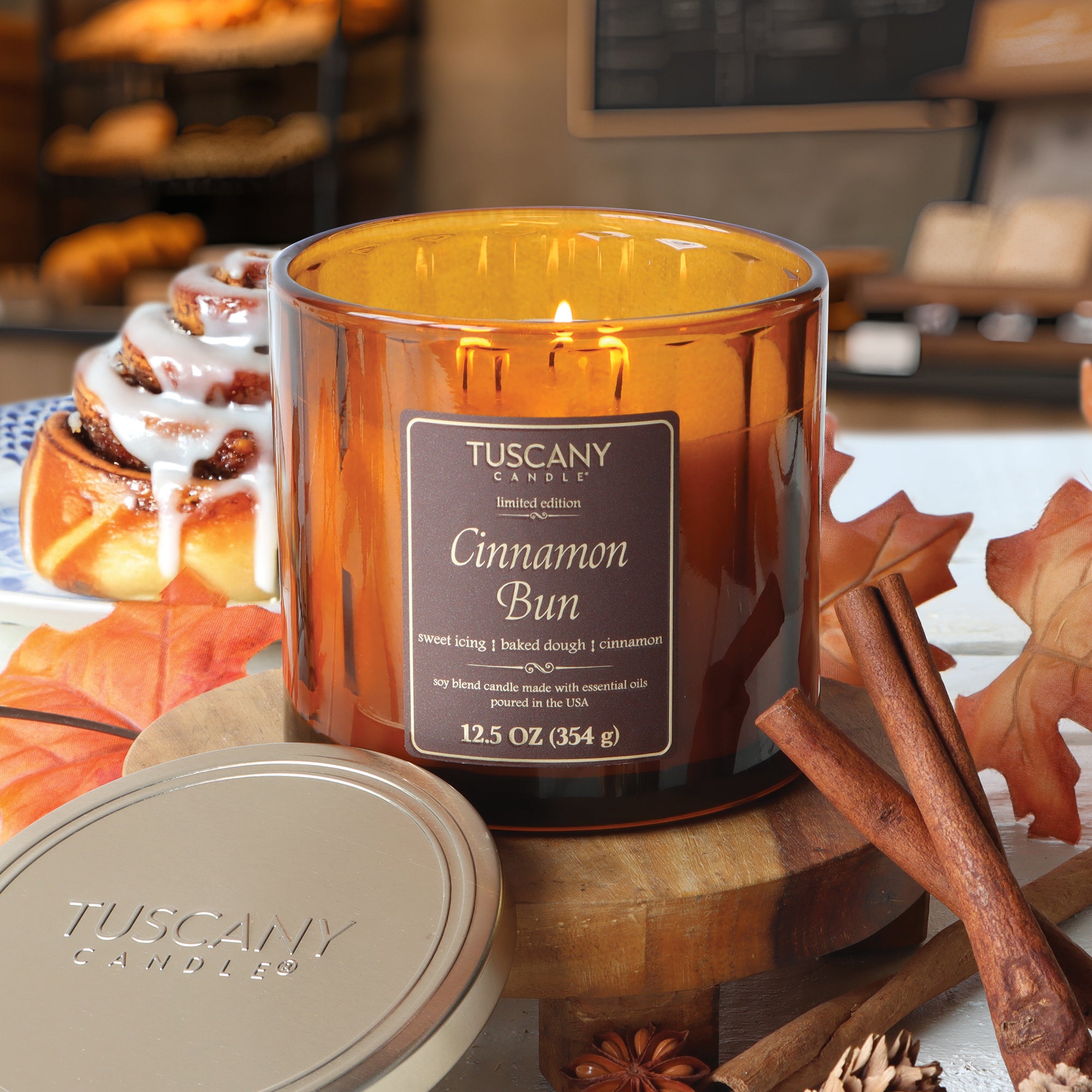 The Tuscany Candle® SEASONAL Cinnamon Bun: Icing & Dough Scented Fall Candle (12.5 oz) is lit on a wooden surface, surrounded by cinnamon sticks, leaves, and a cinnamon roll—capturing the ultimate cozy autumn vibe.