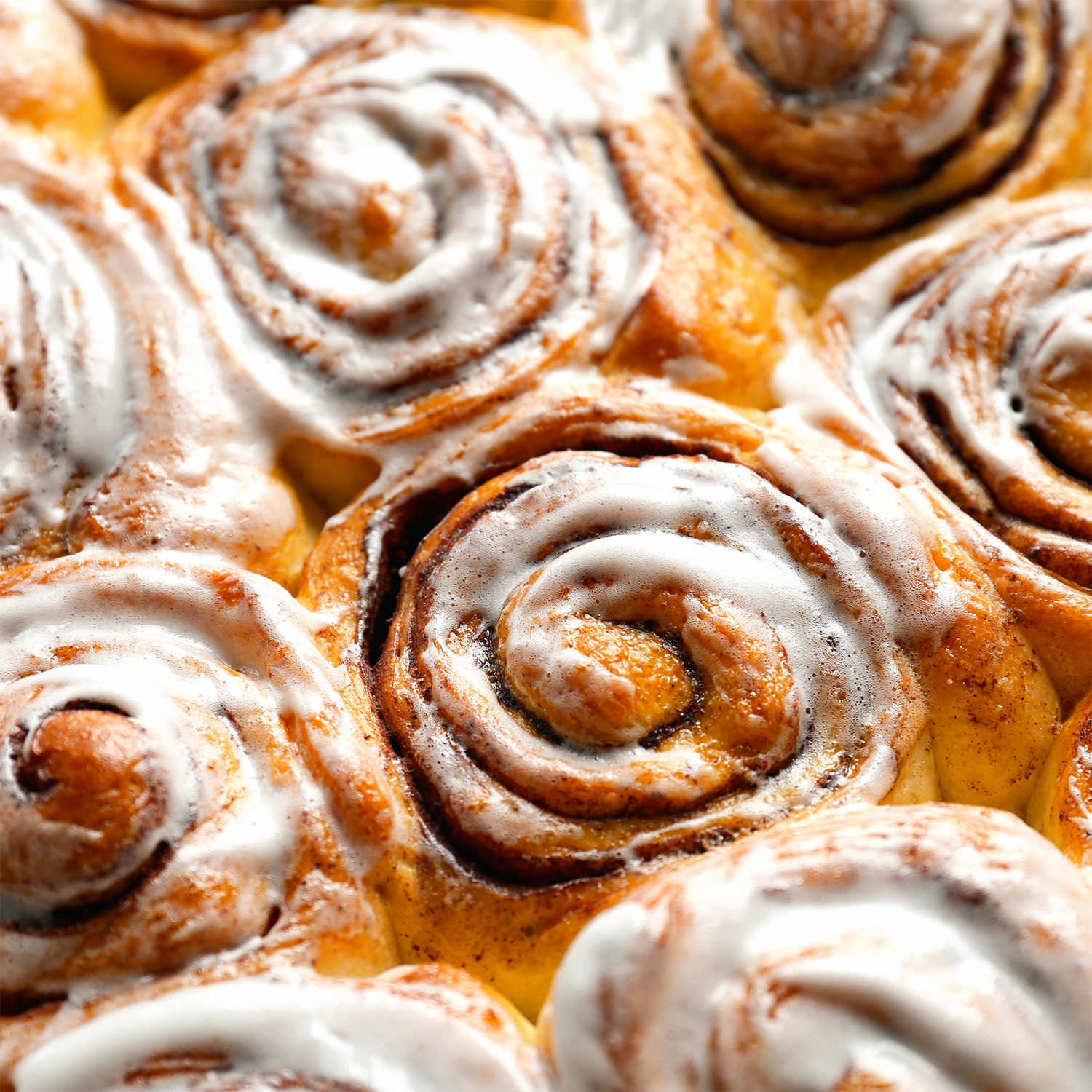 Close-up of freshly baked cinnamon rolls topped with white icing, nestled together in a baking tray—just like the irresistible scent from Tuscany Candle® SEASONAL Cinnamon Bun: Icing & Dough Scented Fall Candle (12.5 oz), made from a soy blend wax to fill your space with cozy fall vibes.