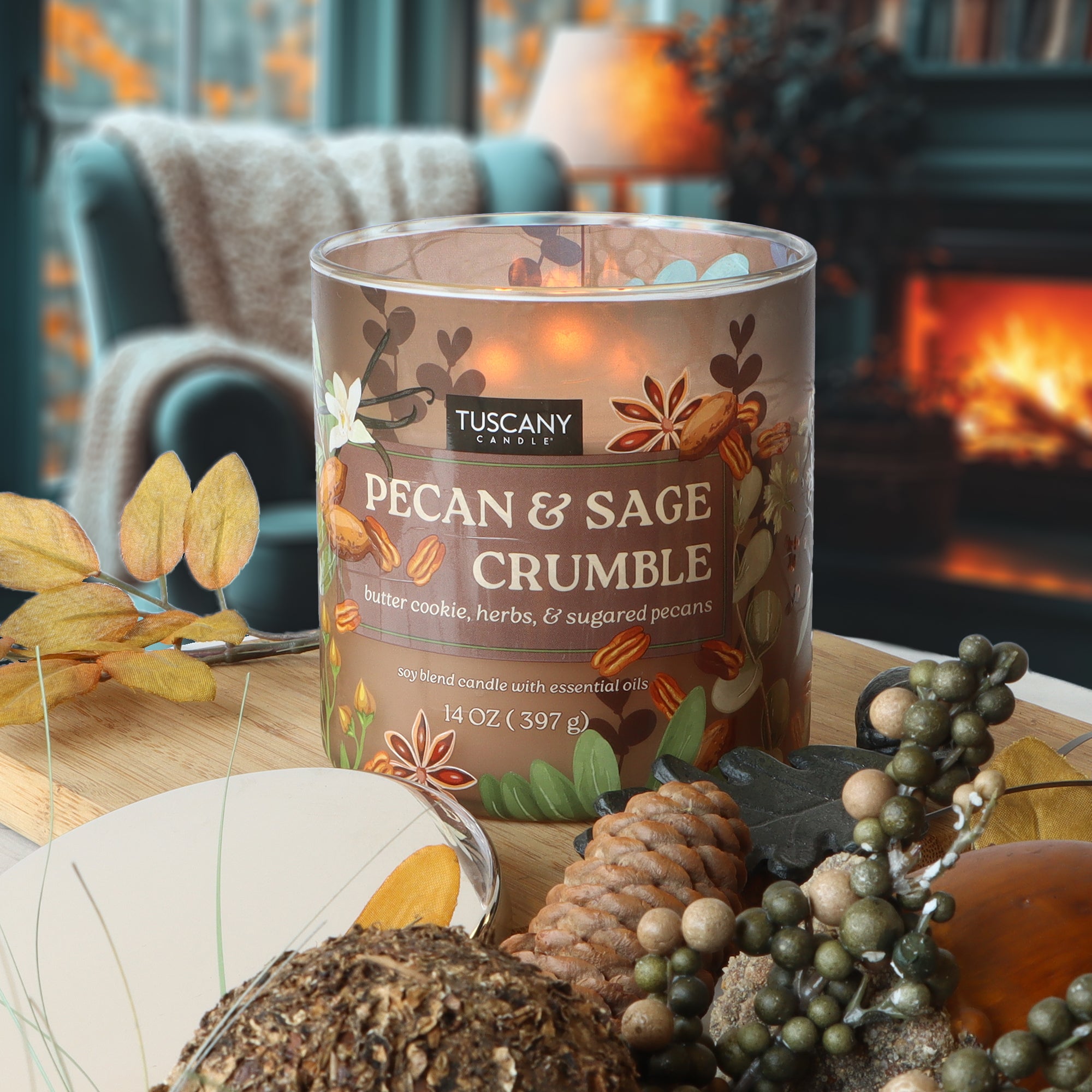 A Tuscany Candle® SEASONAL "Pecan & Sage Crumble: Butter Cookie & Herbs" 14 oz fall candle sits on a table with autumn décor, near a lit fireplace and an armchair draped in a cozy blanket.