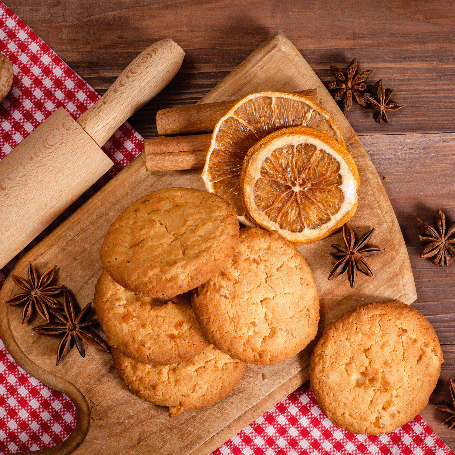 Five Pecan & Sage Crumble: Butter Cookie & Herbs scented candles from Tuscany Candle® SEASONAL rest on a wooden board with dried orange slices, cinnamon sticks, and star anise, capturing the cozy essence of fall home fragrance. A rolling pin and red gingham cloth add a rustic touch to the wooden table setting.