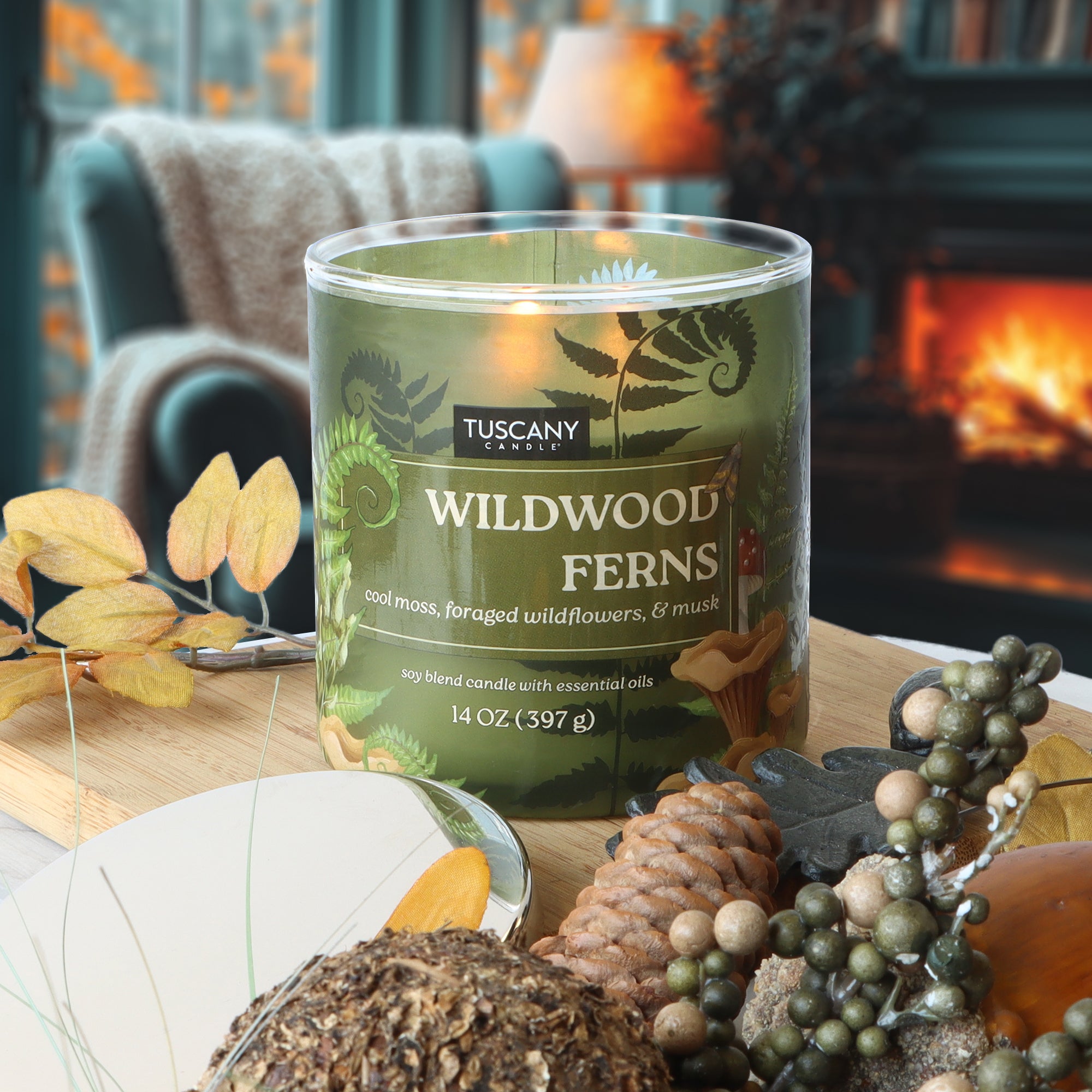 A Tuscany Candle® SEASONAL Wildwood Ferns: Cool Moss & Wildflower Scented Fall Candle (14 oz) sits on a table with autumn decor and pinecones, with a fireplace and armchair in the background.