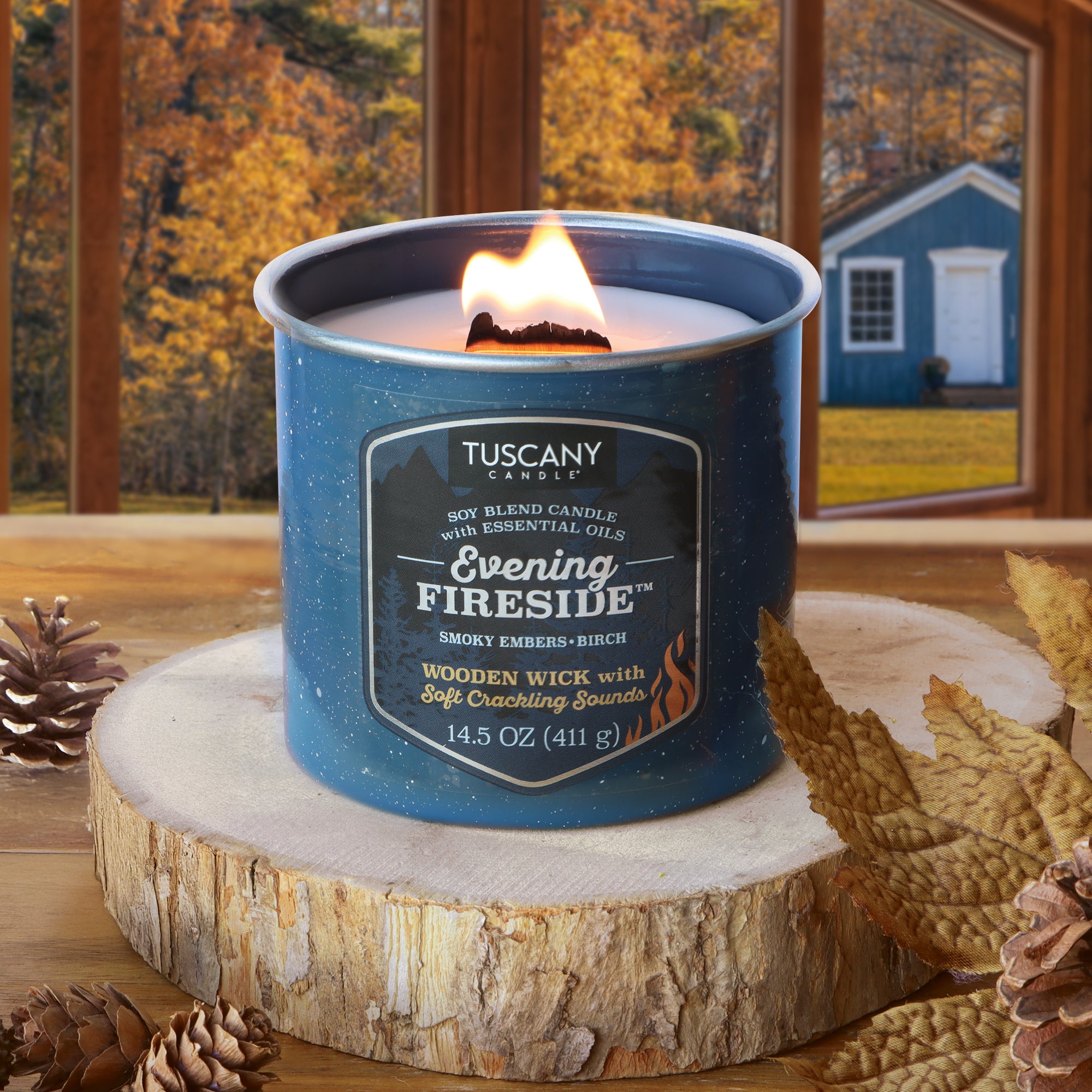 A 14.5 oz Tuscany Candle® SEASONAL Evening Fireside®: Smoky Embers & Birch Scented Fall Wooden Wick Candle glows atop a wooden surface, filling the air with the comforting scent of smoky embers and birch. Surrounded by pinecones and set against a backdrop of autumn trees and a blue house visible through the window, this fall candle creates a cozy, inviting atmosphere reminiscent of crackling fires on chilly evenings.