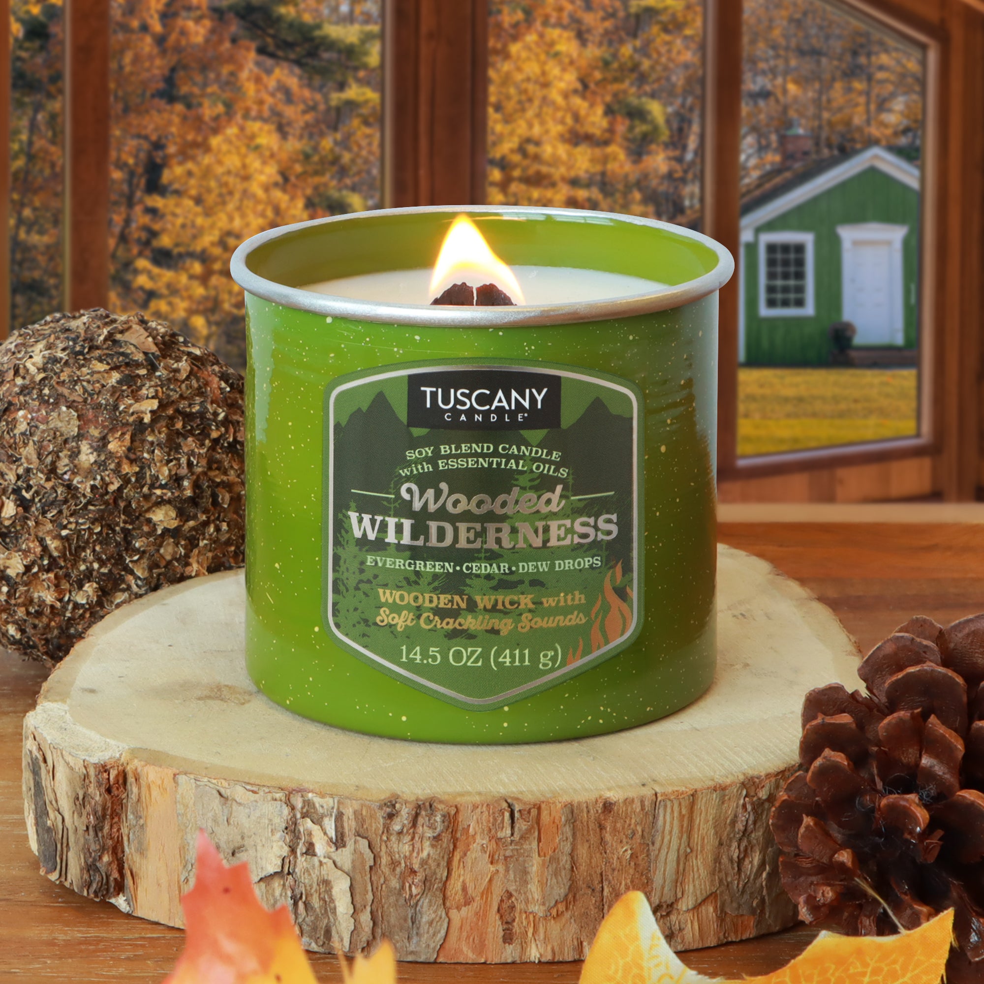 A Tuscany Candle® SEASONAL Wooded Wilderness: Evergreen & Cedar Scented Fall Wooden Wick Candle (14.5 oz) burns on a wooden surface with pinecones, a log, and autumn trees visible through the window in back.