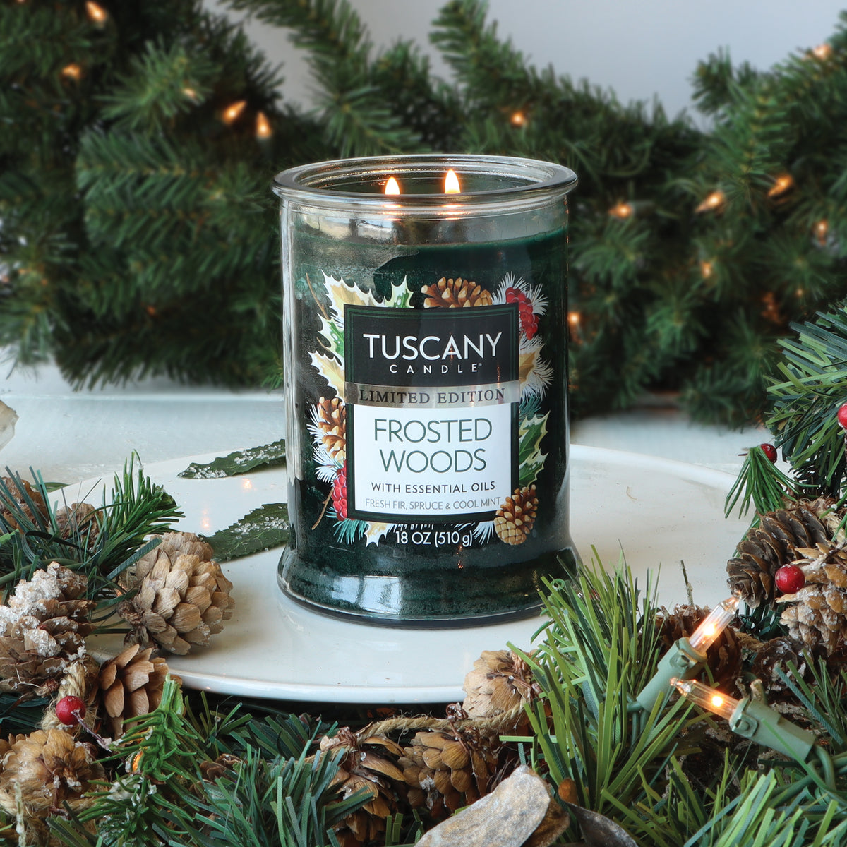 Tuscany Candle® SEASONAL Frosted Woods: Fresh Fir & Spruce Scented Holiday Candle (18 oz) in a two-wick jar with a dark green label, surrounded by pine branches, pinecones, and string lights.