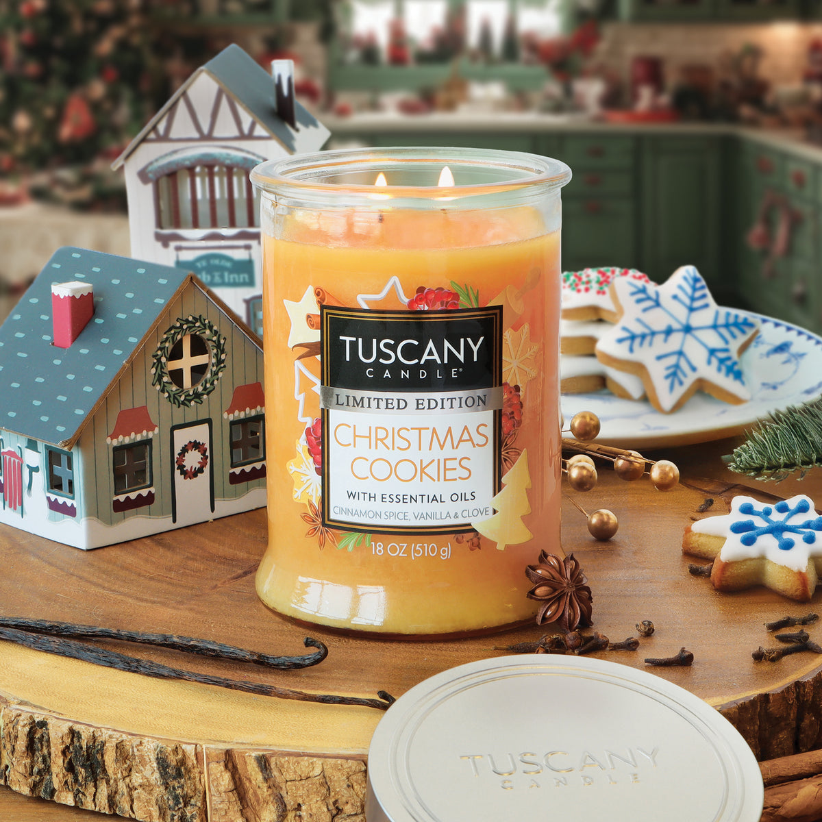 A Tuscany Candle® SEASONAL Christmas Cookies: Cinnamon Spice & Vanilla Scented Holiday Candle (18 oz) with orange wax, sitting on a wooden board next to snowflake cookies, house figurines, anise, and cloves.