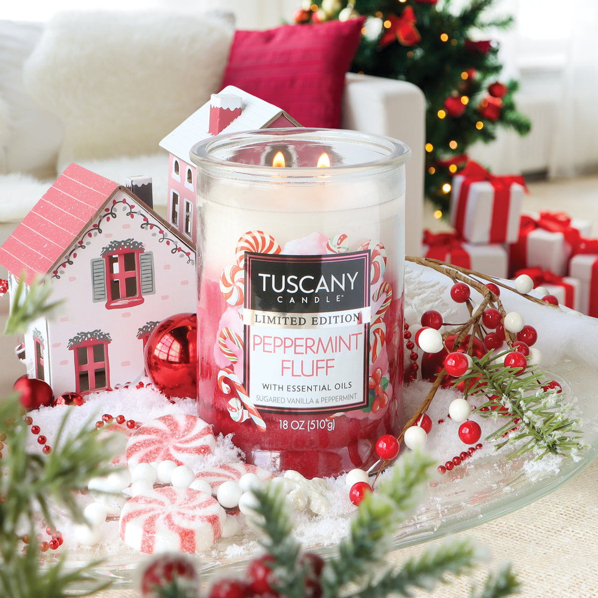 A Peppermint Fluff: Sugared Vanilla & Peppermint Scented Holiday Candle (18 oz) by Tuscany Candle® SEASONAL in a clear glass jar on a tray with faux snow, miniature houses, red ornaments, and candies.