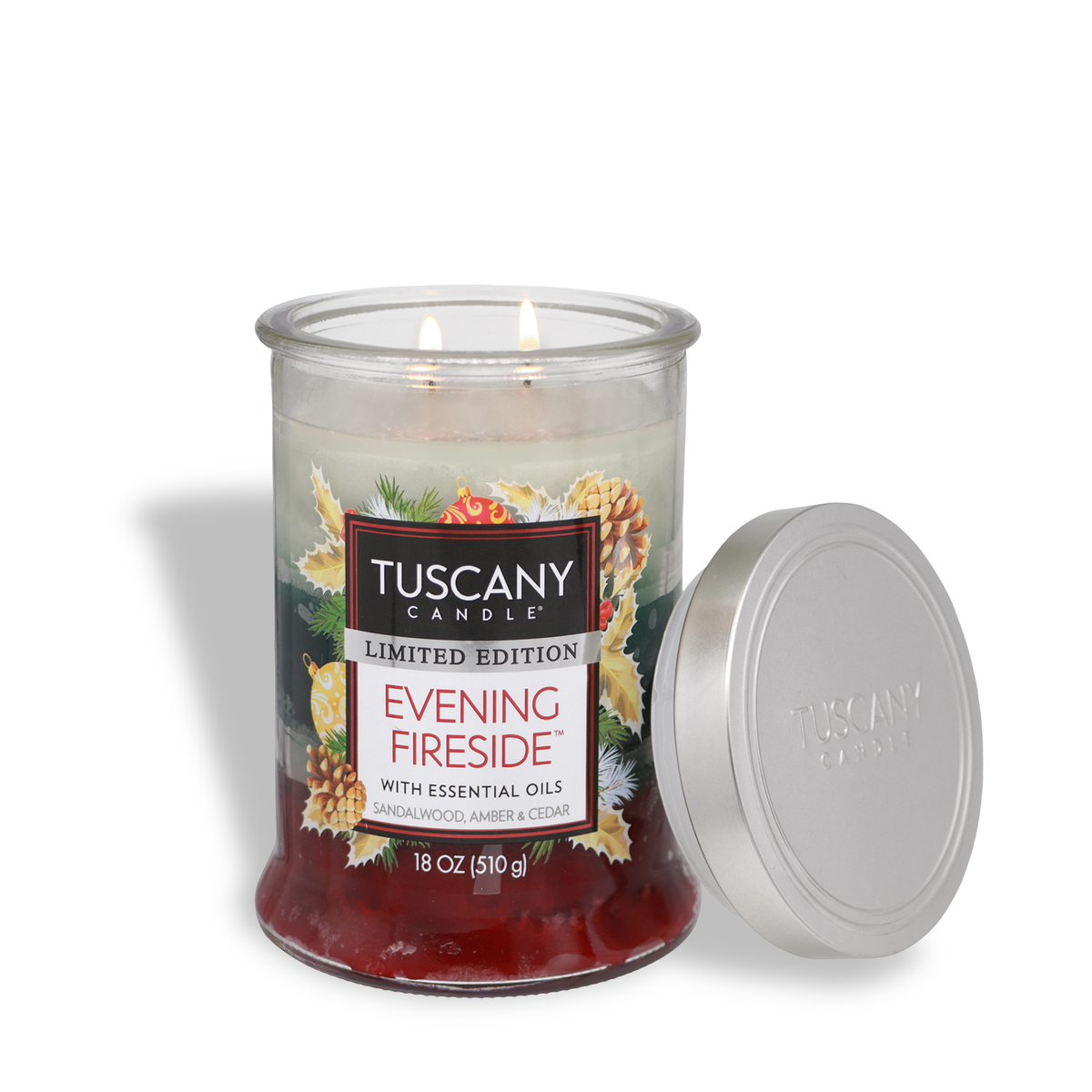 A two-wick Evening Fireside®: Sandalwood & Amber Scented Holiday Candle (18 oz) in a glass jar with a festive label and its silver lid resting beside it.