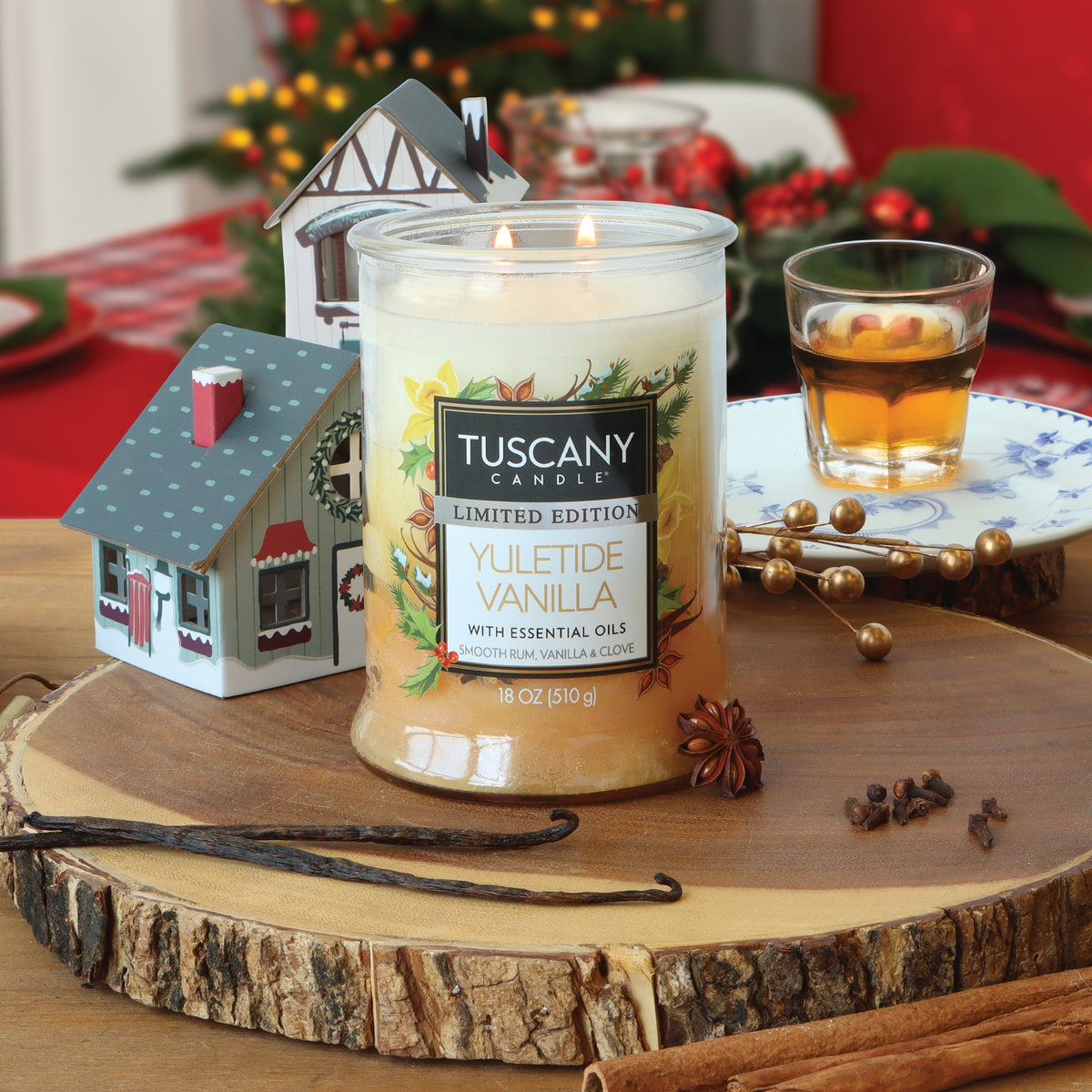 A Tuscany Candle® SEASONAL Yuletide Vanilla: Smooth Rum & Clove Scented Holiday Candle (18 oz) in a two-wick glass jar sitting on a wooden slab, surrounded by vanilla beans, cloves, and miniature house decorations.