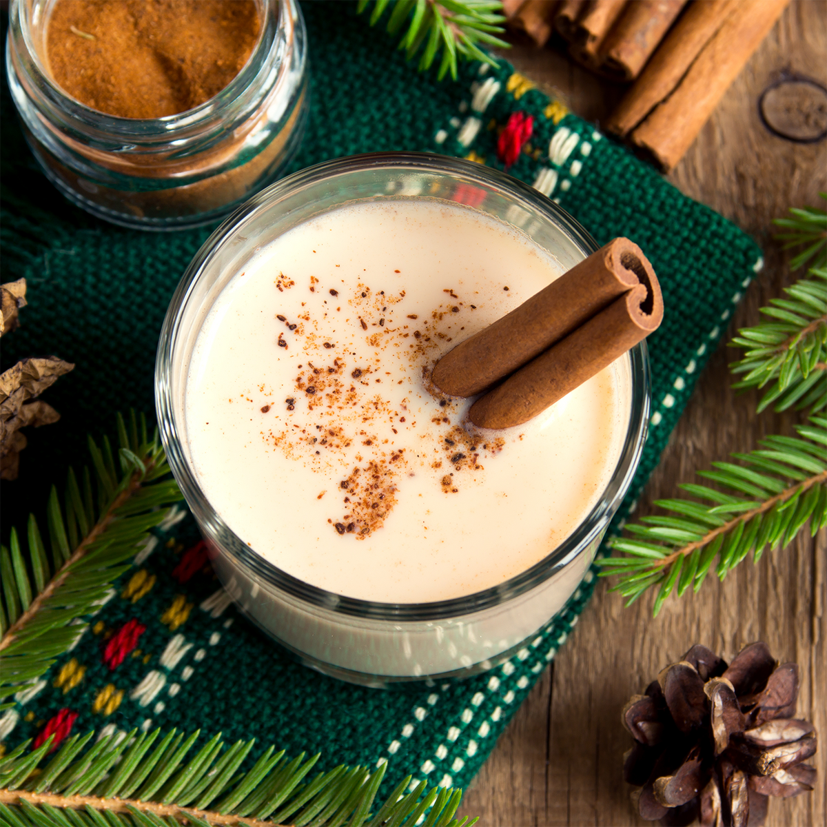 A Yuletide Vanilla: Smooth Rum & Clove Scented Holiday Candle (18 oz) by Tuscany Candle® SEASONAL sits as a glass of eggnog topped with cinnamon sticks, surrounded by pine branches and spices.