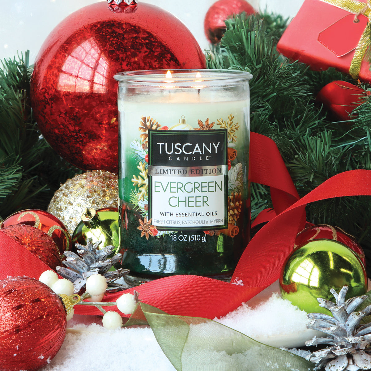 Evergreen Cheer: Fresh Citrus & Patchouli Scented Holiday Candle (18 oz) in a two-wick glass jar, surrounded by red and green Christmas ornaments, pine branches, and ribbon on a snowy surface.