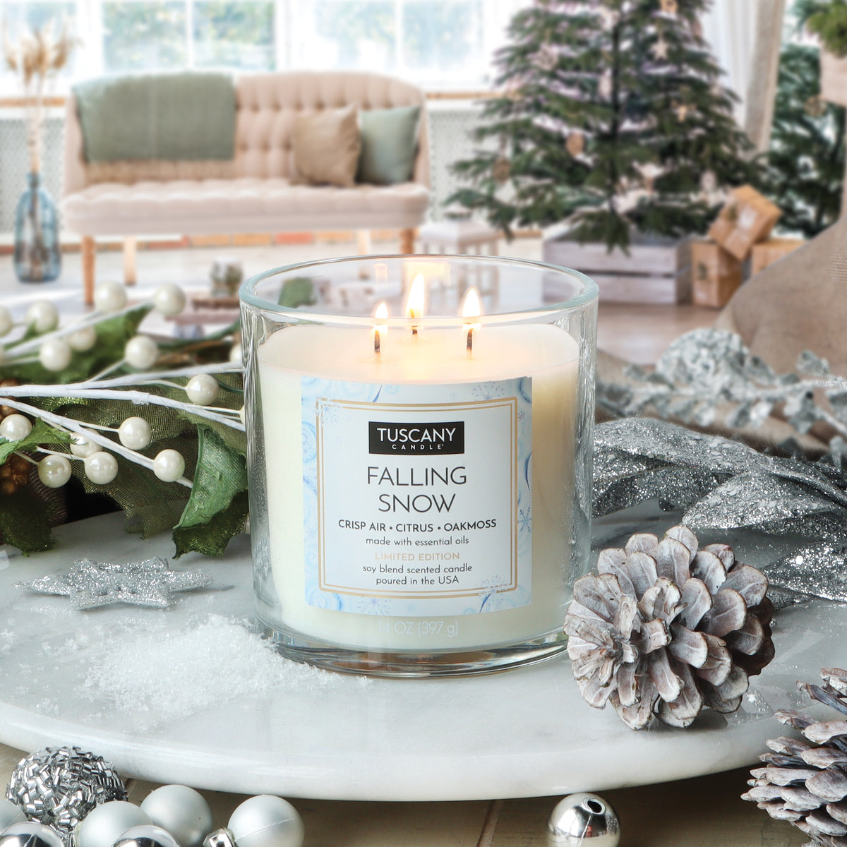 A lit Tuscany Candle® SEASONAL Falling Snow: Crisp Air & Citrus Scented Holiday Candle (14 oz) in a glass jar on a marble surface, surrounded by pinecones, silver ornaments, and snowy decorations.