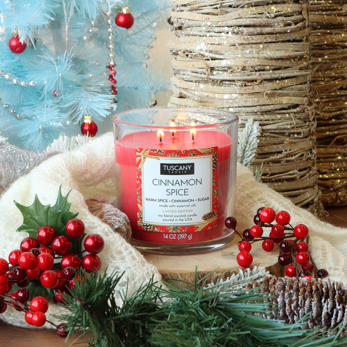 A red Tuscany Candle® SEASONAL Cinnamon Spice: Warm Spice & Sugar Scented Holiday Candle (14 oz) in a glass jar, decorated with holly berries, pine branches, and a white knit wrap.