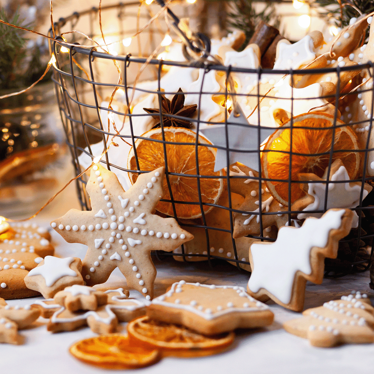 A wire basket filled with star-shaped cookies, dried orange slices, and star anise, surrounded by fairy lights and a Mrs. Claus’s Cookies: Almond & Cardamom Scented Holiday Candle (14 oz).