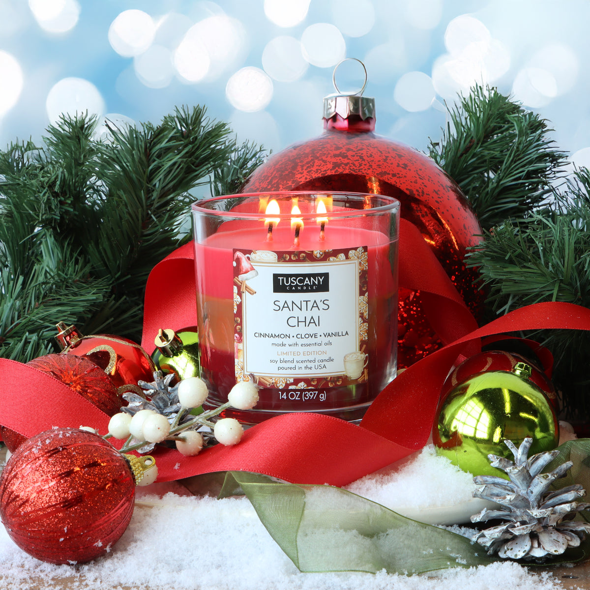 A 14 oz Tuscany Candle® SEASONAL Santa’s Chai: Cinnamon & Clove Scented Holiday Candle glows amidst holiday ornaments, red ribbon, and pine branches on a snowy surface.