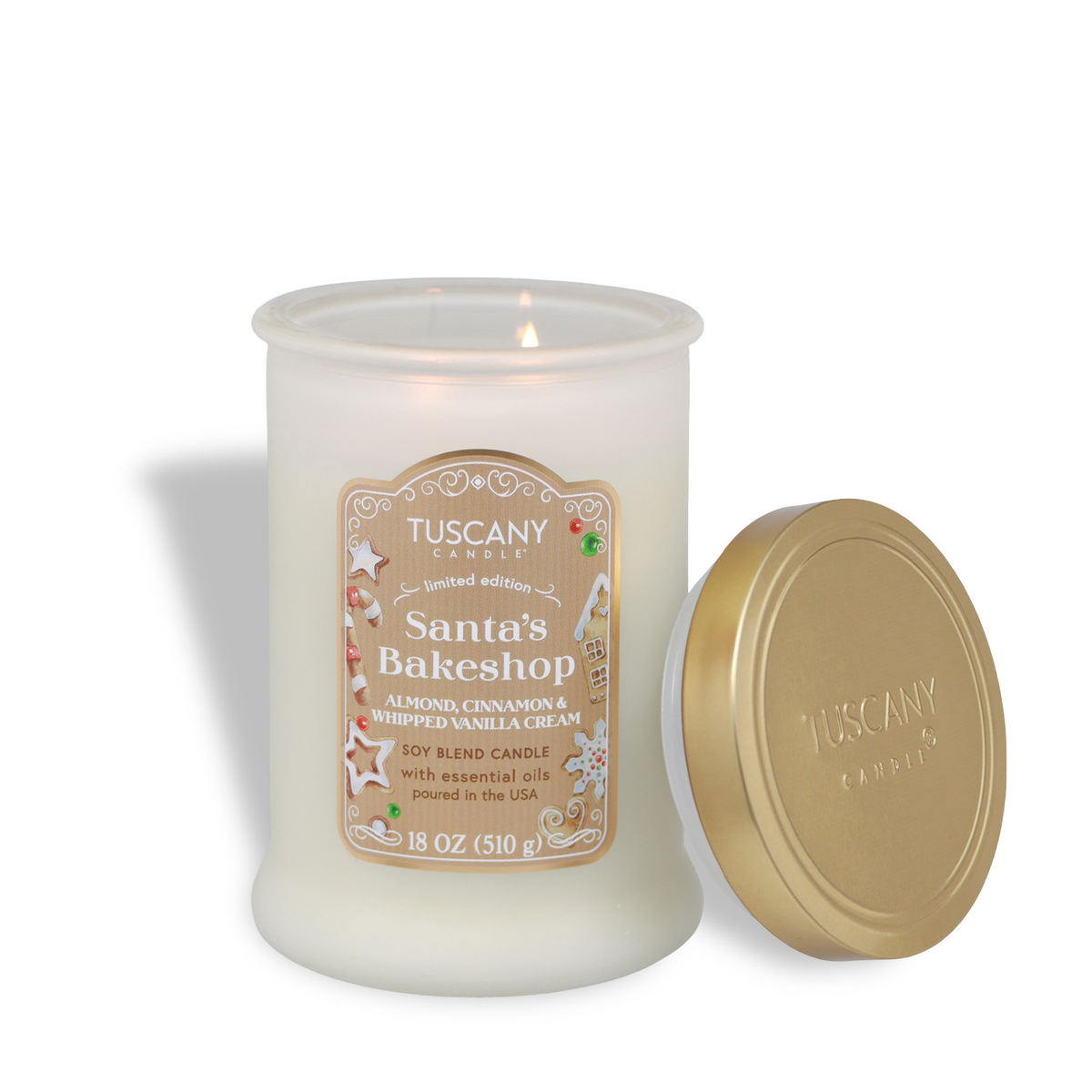 A lit Santa’s Bakeshop: Almond & Vanilla Scented Holiday Candle (18 oz) in a frosted glass jar with a Christmas cookie label, next to its gold lid.