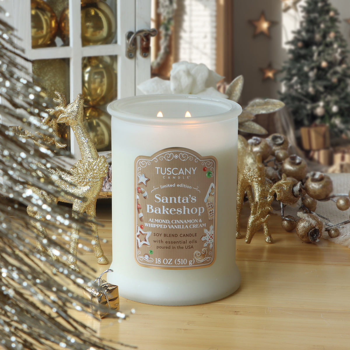 A two-wick Santa’s Bakeshop: Almond & Vanilla Scented Holiday Candle (18 oz) in a frosted glass jar, surrounded by gold reindeer ornaments and holiday decorations.