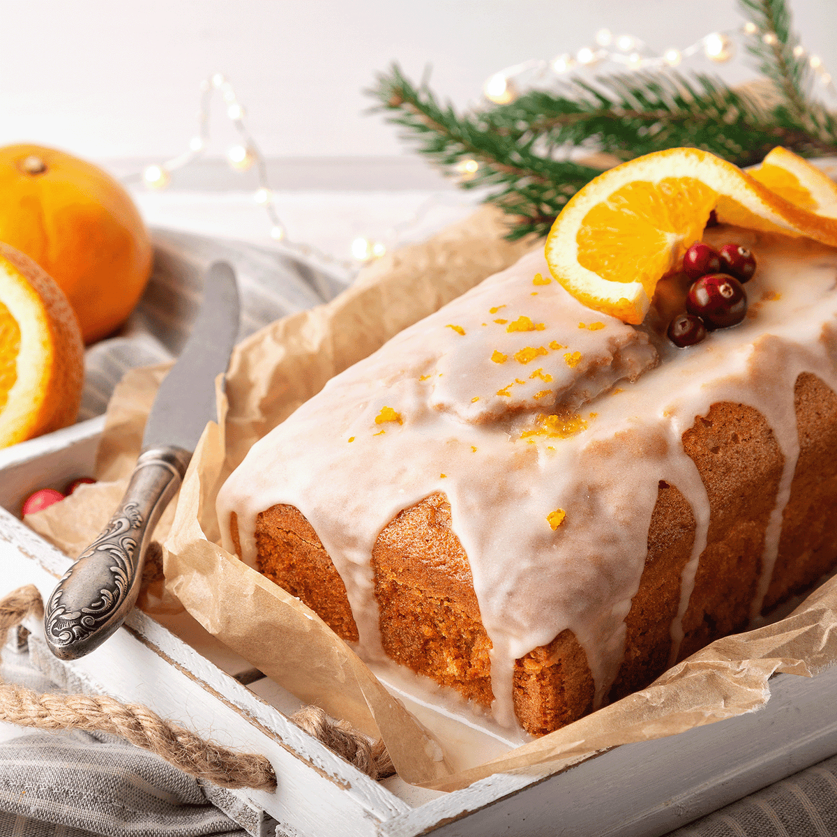 A glazed loaf cake topped with orange slices and cranberries, served near a Gingersnap Citrus: Candied Orange & Cinnamon Scented Holiday Candle (18 oz) on a white wooden tray.