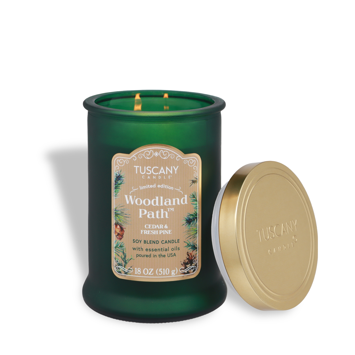 A lit two-wick Woodland Path®: Cedar & Pine Scented Holiday Candle (18 oz) in a green glass jar with a tan label and a gold lid.
