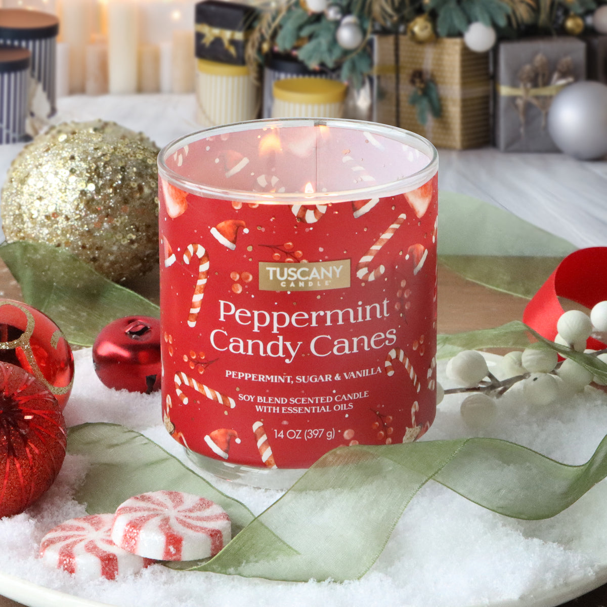 A 14 oz Tuscany Candle® SEASONAL Peppermint Candy Canes: Sugar & Vanilla Scented Holiday Candle with a red label featuring candy canes and Santa hats, on fake snow with ornaments and peppermint candies.