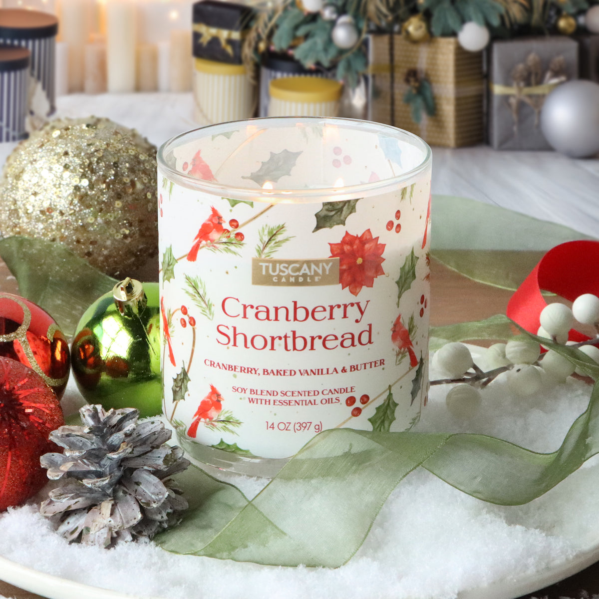 A Tuscany Candle® SEASONAL Cranberry Shortbread: Baked Vanilla & Butter Scented Holiday Candle (14 oz) in a glass jar with a cranberry and poinsettia print, sitting on fake snow, ornaments, and ribbons.